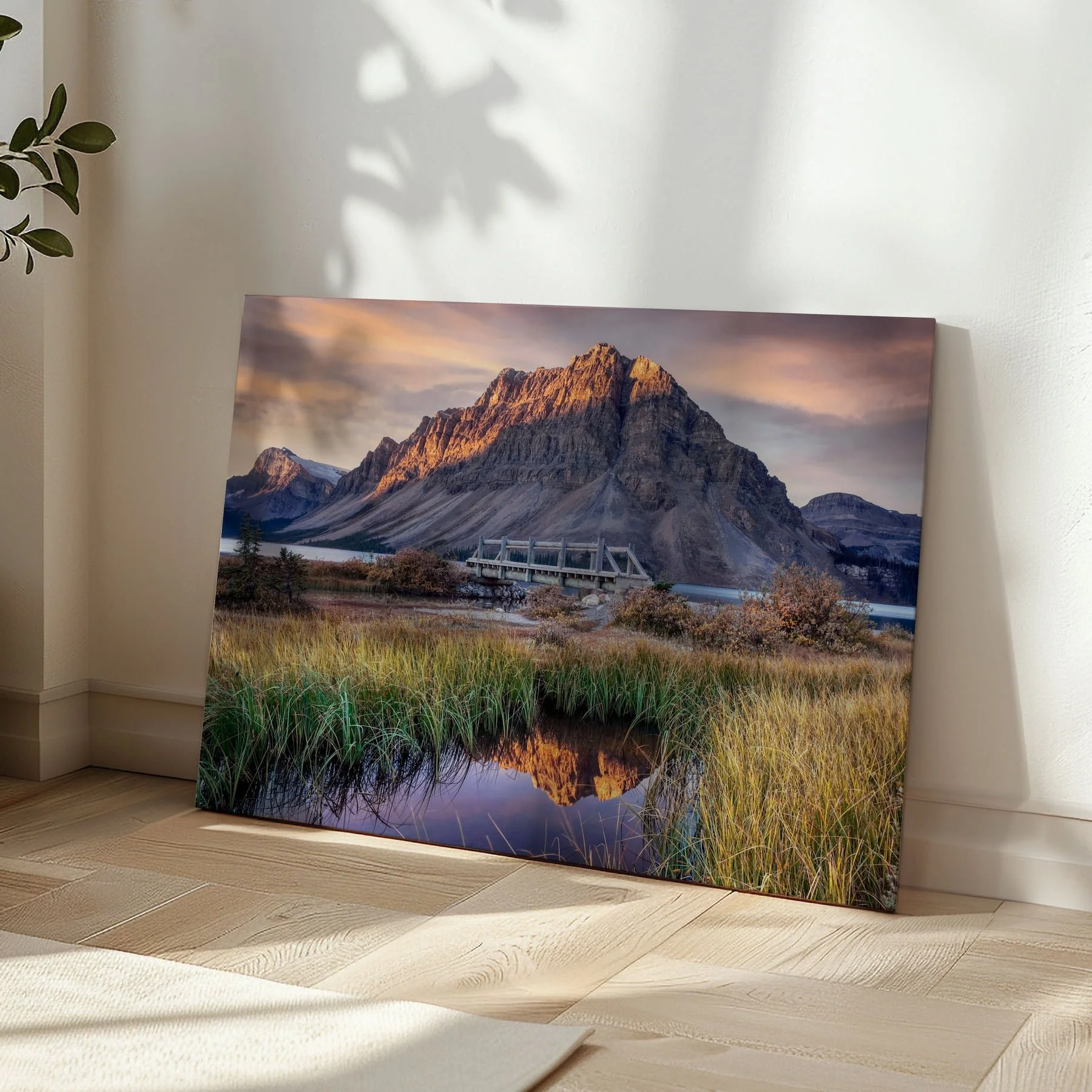 Bow Lake at Dawn gallery-wrapped canvas print leaning in a sunlit room corner on herringbone hardwood flooring, with soft leaf shadows on the wall beside it.
