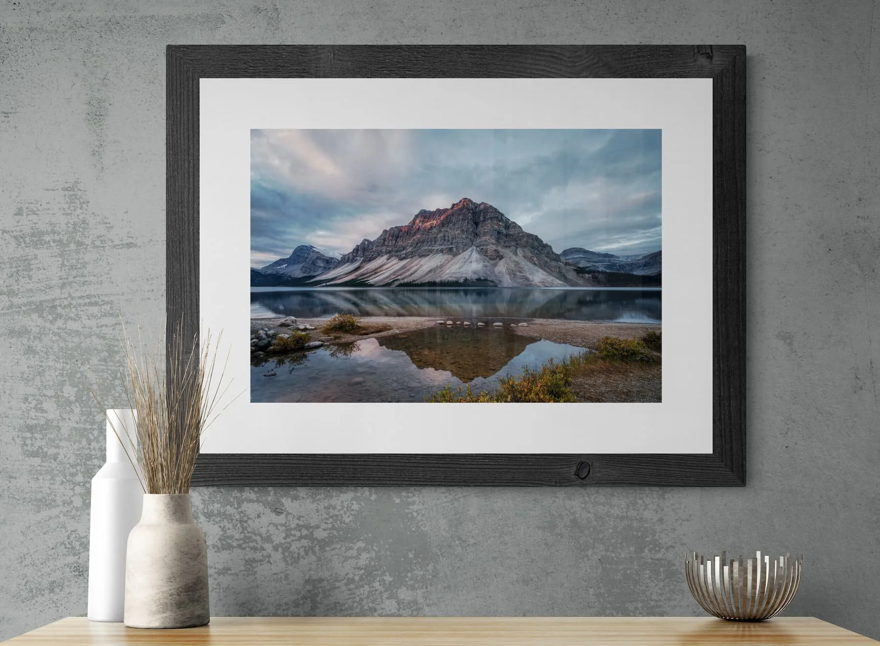 Bow Lake Reflections photograph in a wide dark barnwood frame with white mat, mounted on an industrial concrete wall above a wooden sideboard with ceramic vases and dried grass.