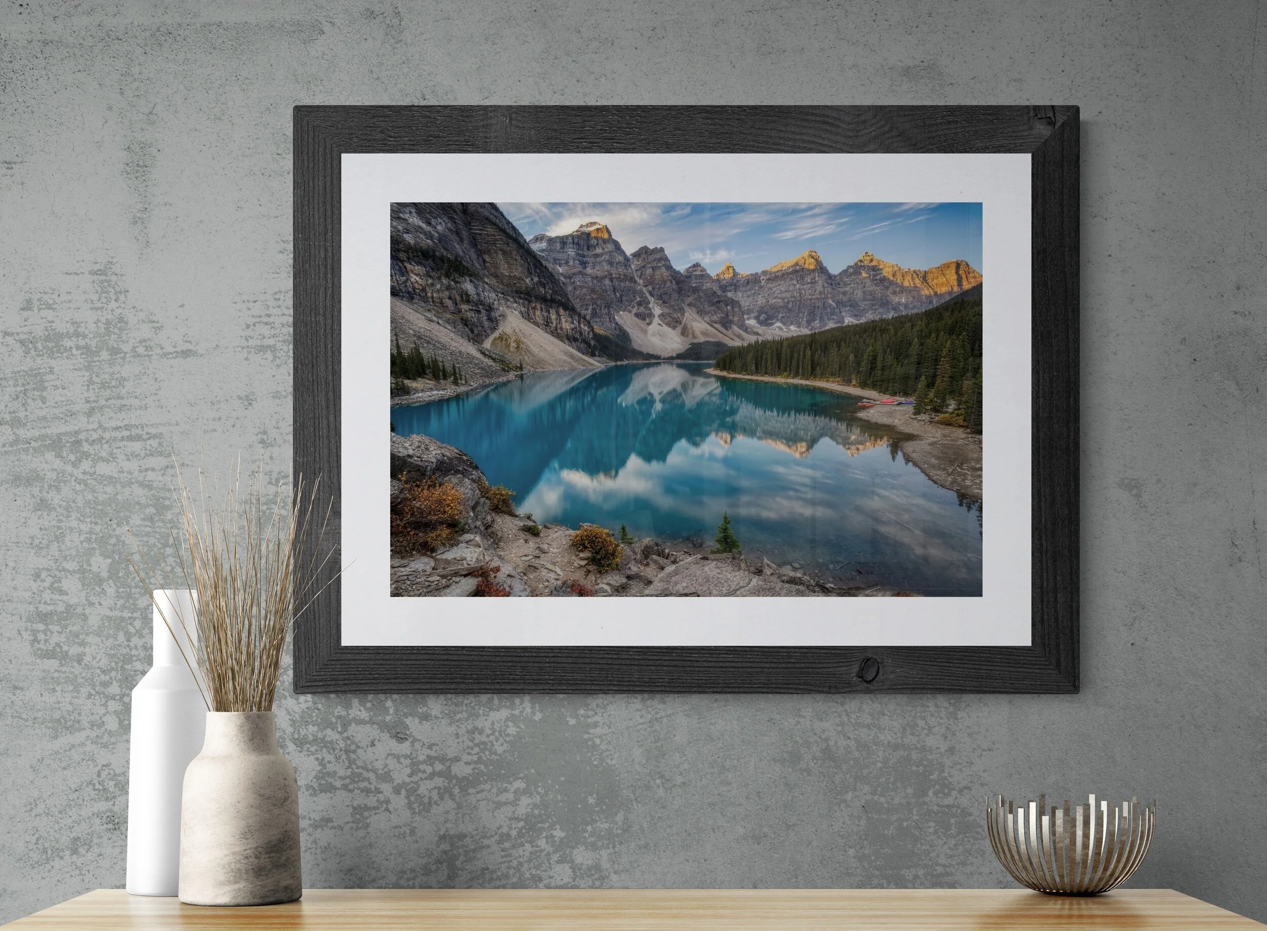 Moraine Lake Sunrise photograph in a wide dark barnwood frame with white mat, mounted on an industrial concrete wall above a wooden sideboard with ceramic vases and dried grass.