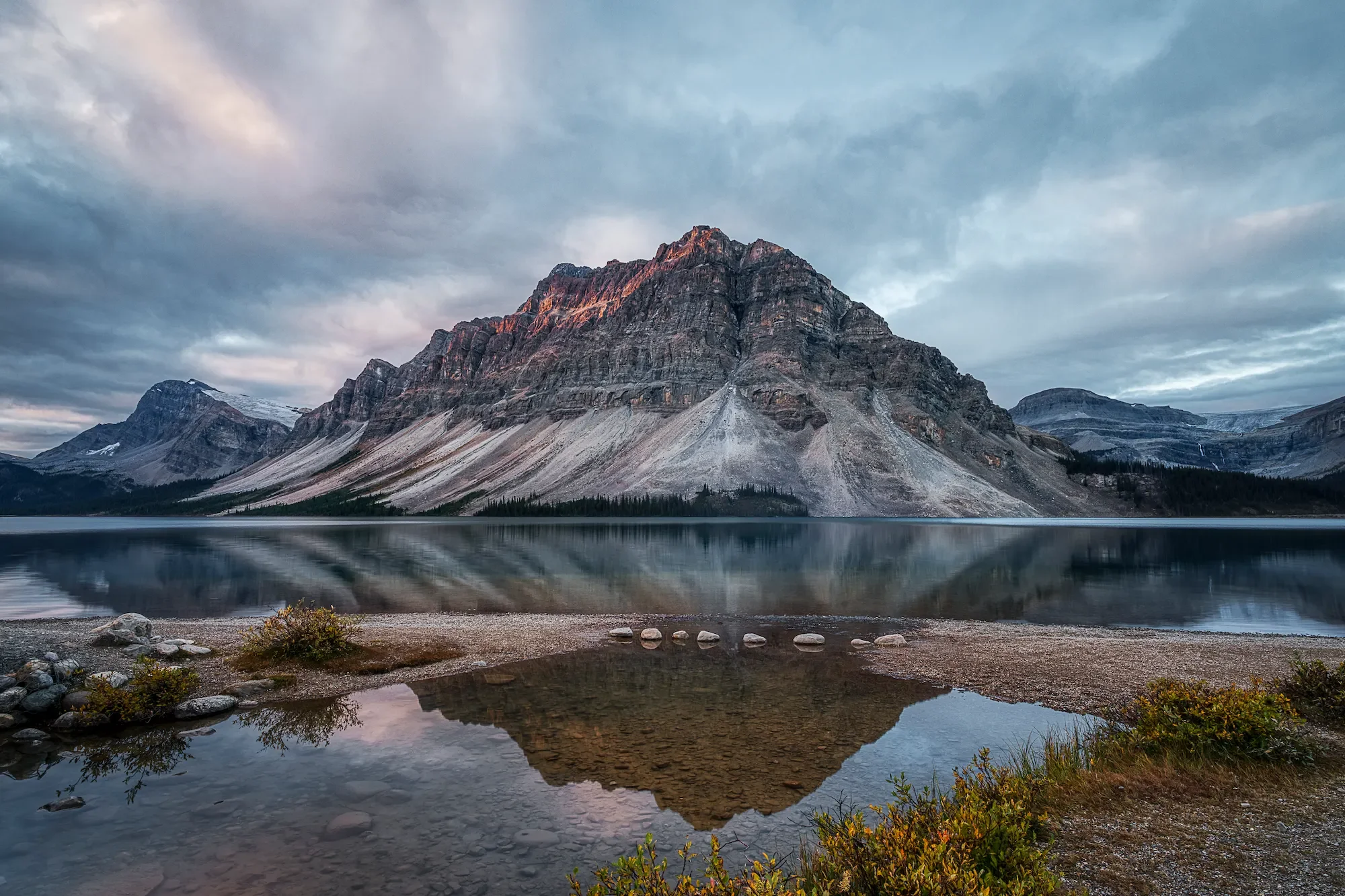 A mountain reflected in a calm lake with rocks and bushes in the foreground and a cloudy sky overhead.