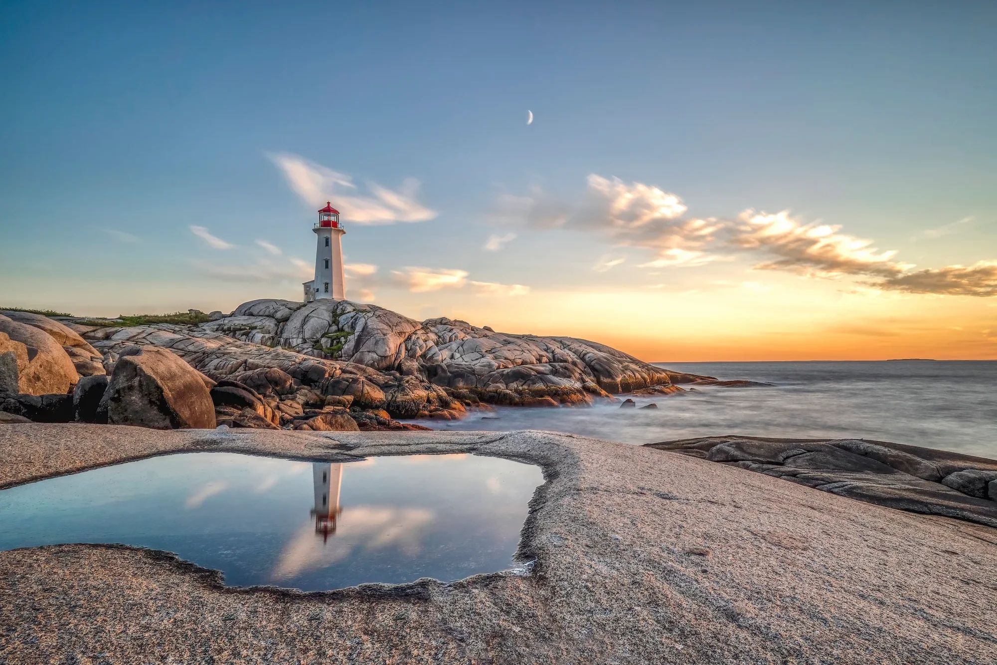 Peggy's Cove Lighthouse on rocky terrain during sunset, with a reflection of the lighthouse and sky in a small pool of water on the sandy ground.