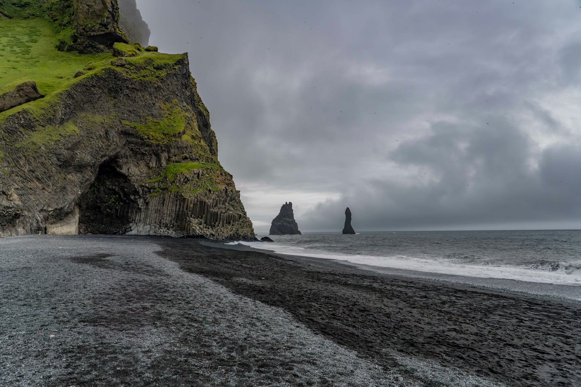 Black sand beach, basalt cliffs and Reynisdrangar sea stacks beneath a cloudy sky at Reynisfjara, Iceland.