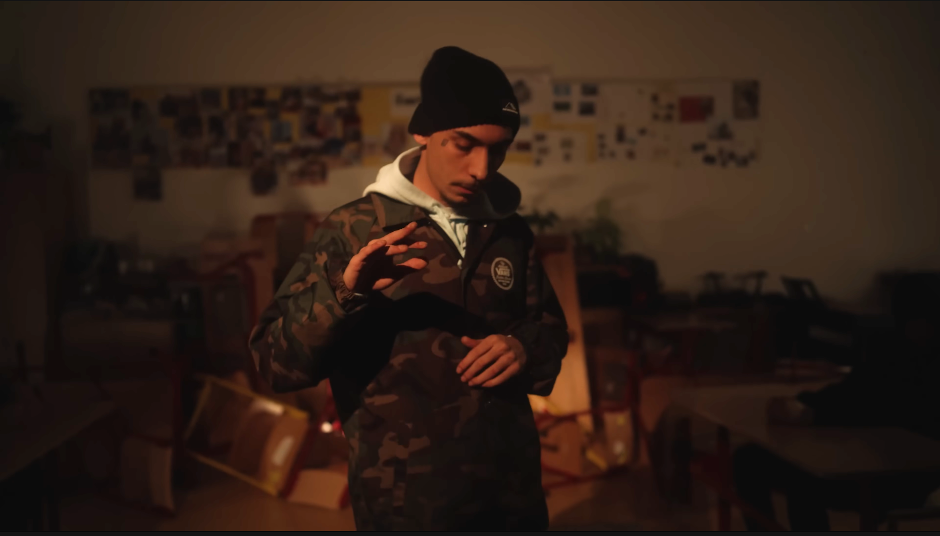 Young man in camouflage jacket and black beanie in a dimly lit room, looking down thoughtfully.