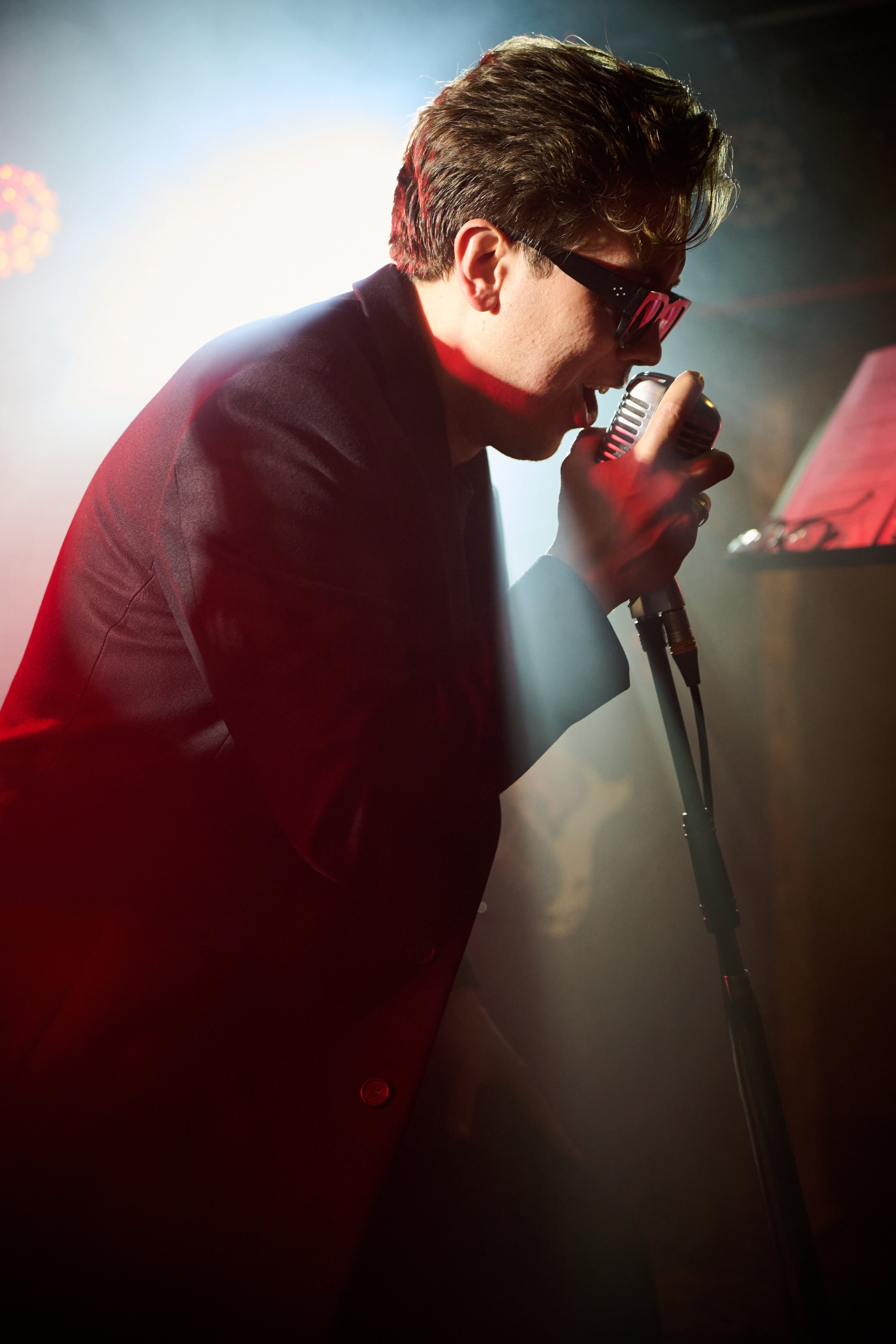 Young man singing into a microphone on stage with colorful lighting and music stand in the background.