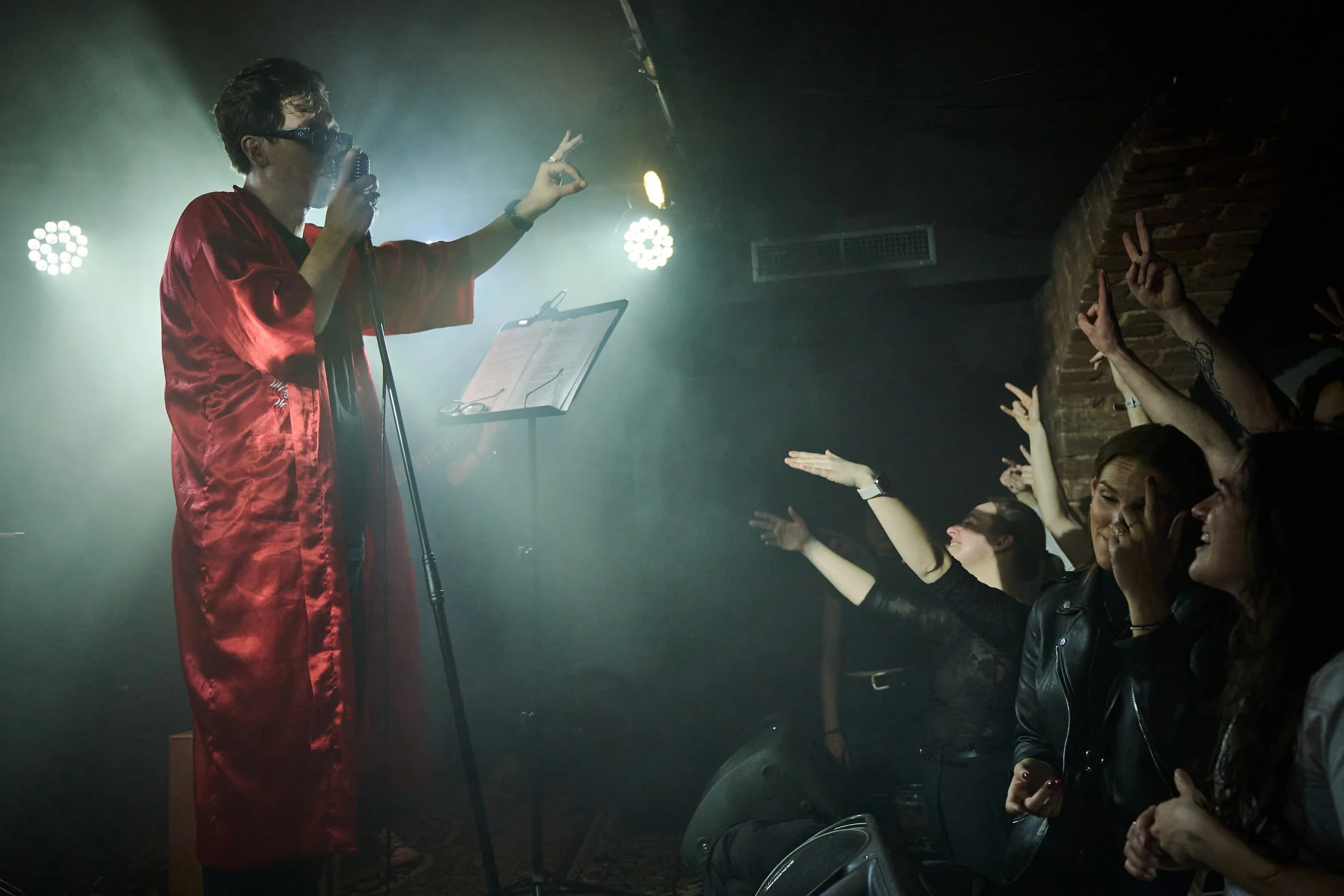 Performer in red satin robe singing into a vintage microphone on stage, with an audience reaching out and making peace signs.