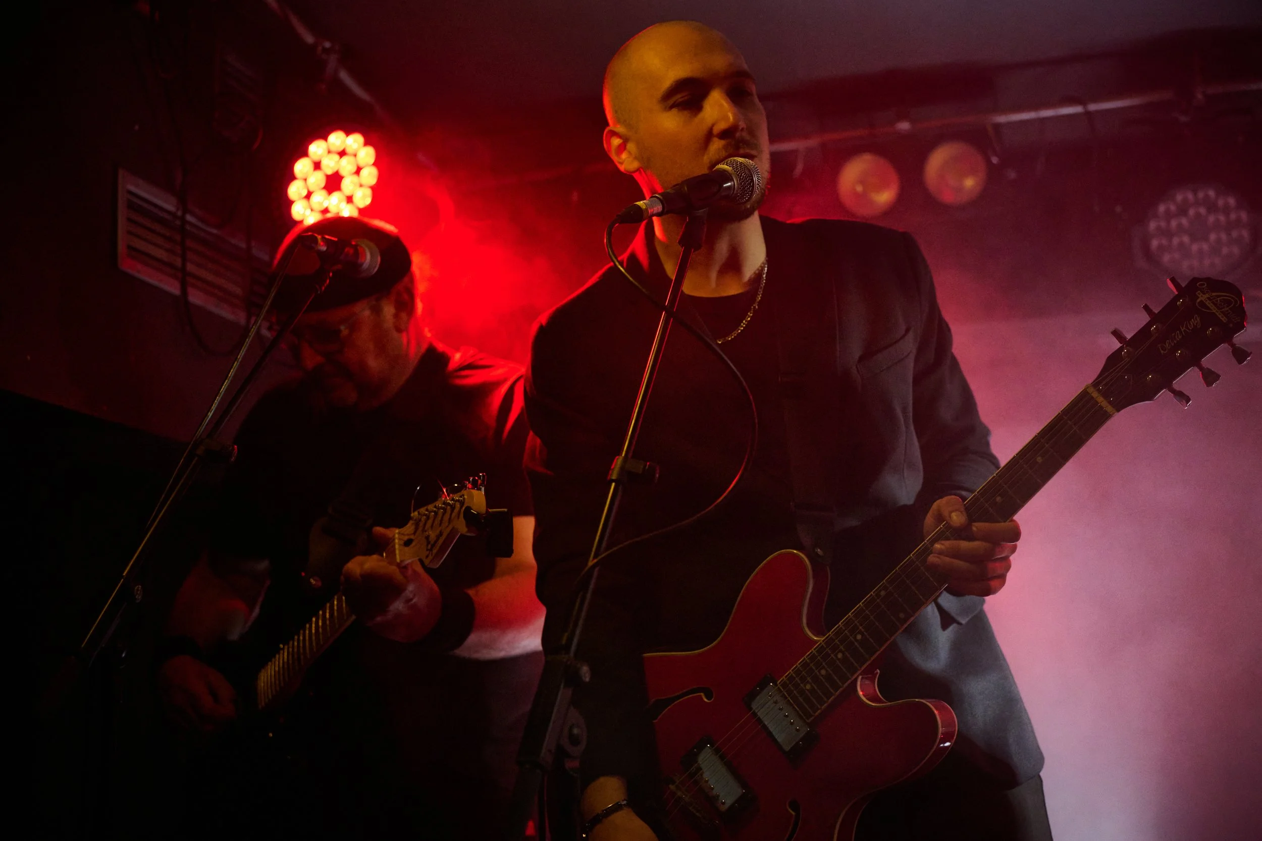 Two musicians performing on stage with red lighting; one playing an electric guitar and singing into a microphone, and the other playing a different string instrument in the background.