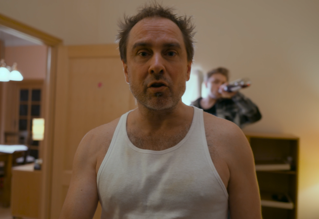 A middle-aged man with disheveled hair, wearing a white tank top, looking directly at the camera in a room, with a person in the background holding a beer bottle.