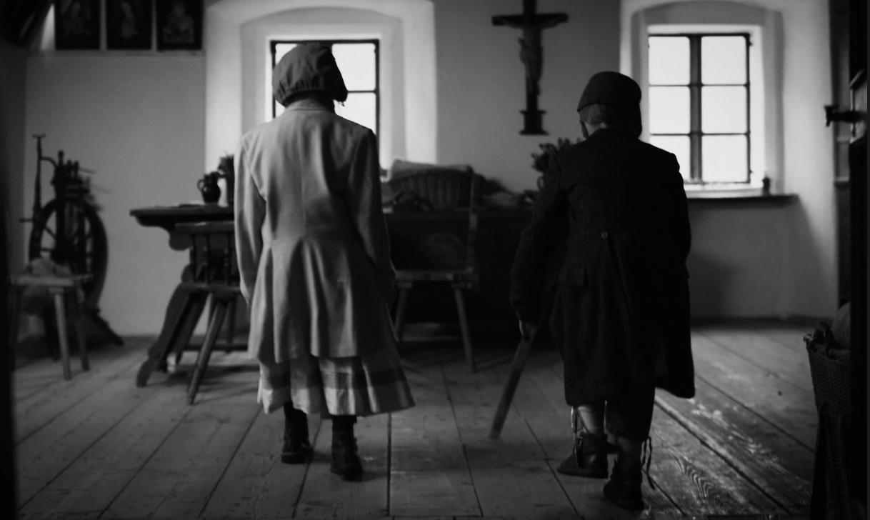 Two children dressed in vintage clothing standing inside a historical room with wooden floors and simple furnishings, seen from behind.