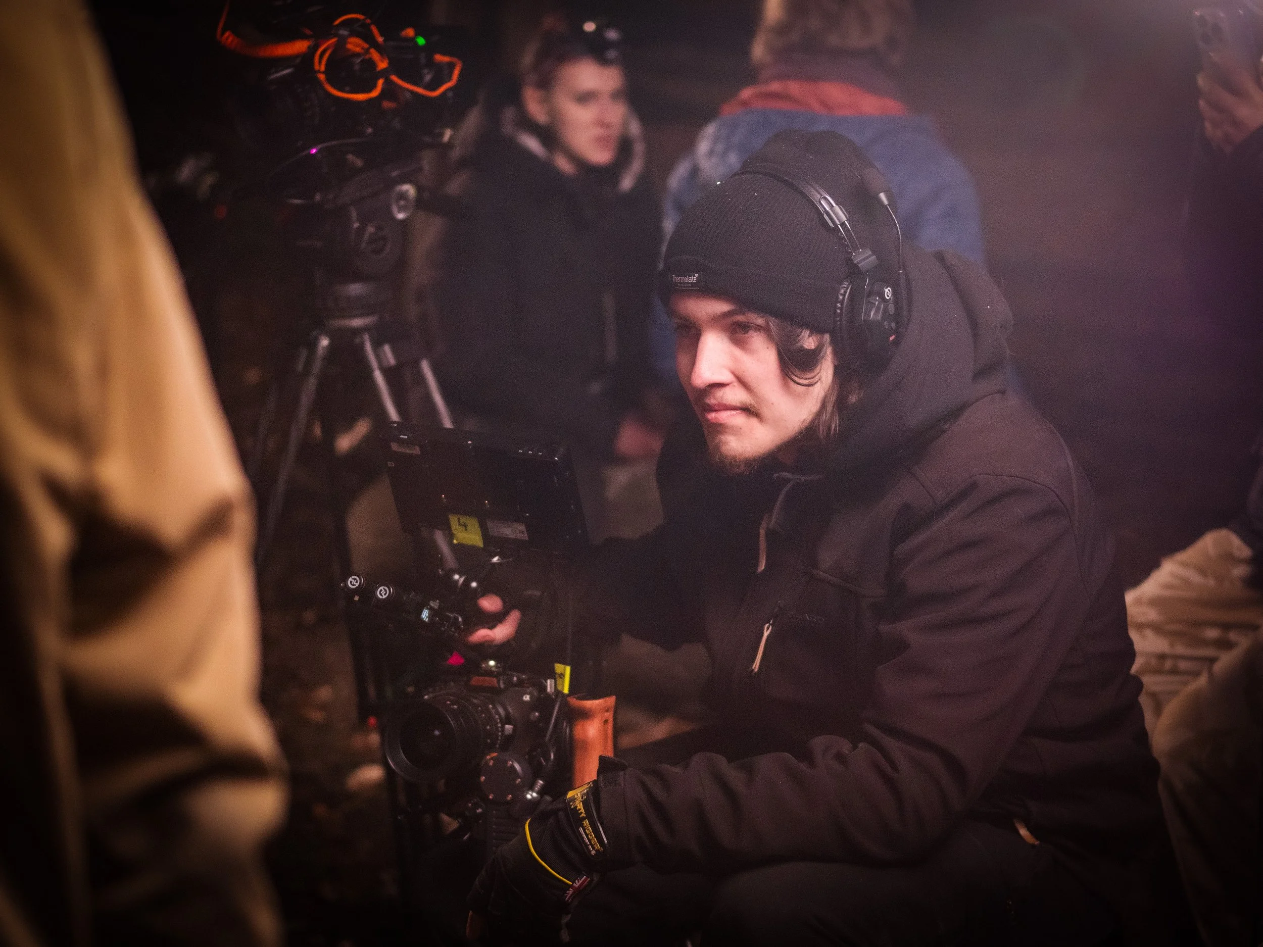 People gathered around a camera and filming equipment in a low-light setting, some wearing headsets, indicating a filming or production environment.
