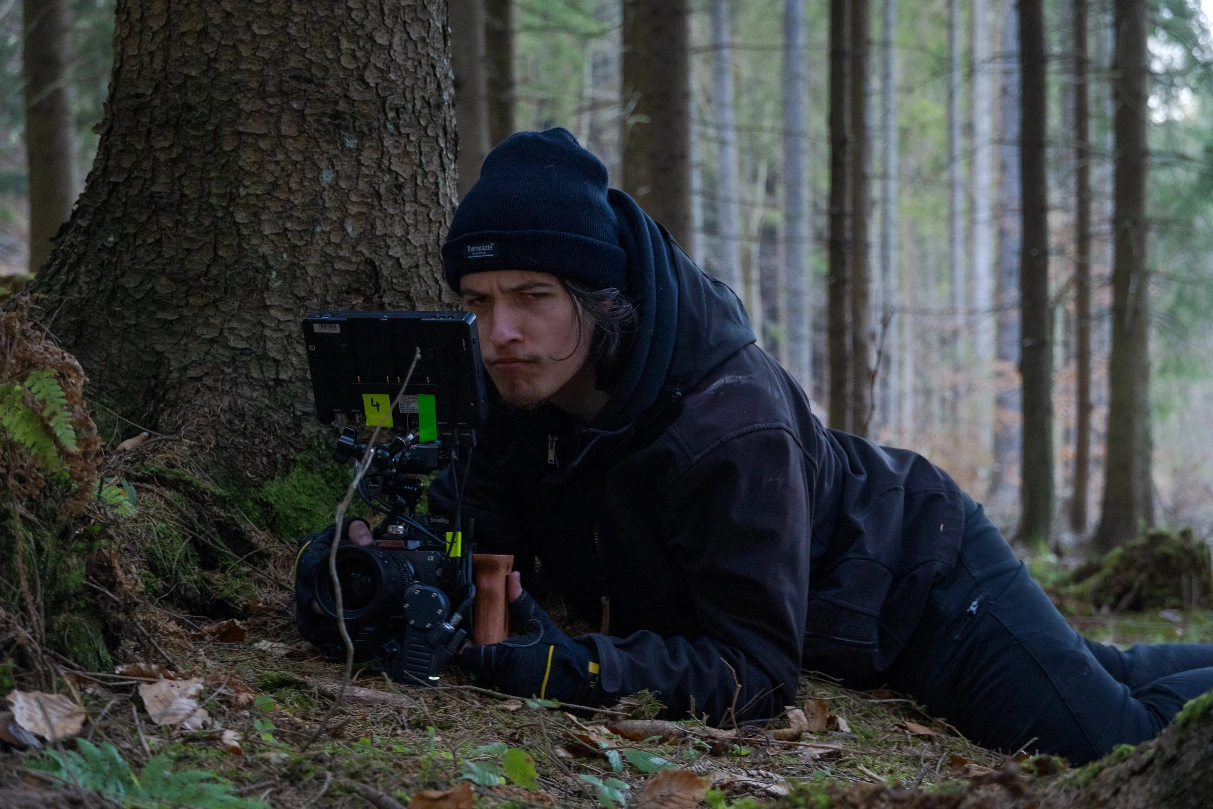 A person wearing a beanie and black clothing is lying on the ground in a forest, examining a piece of equipment mounted on a tripod near a tree trunk.