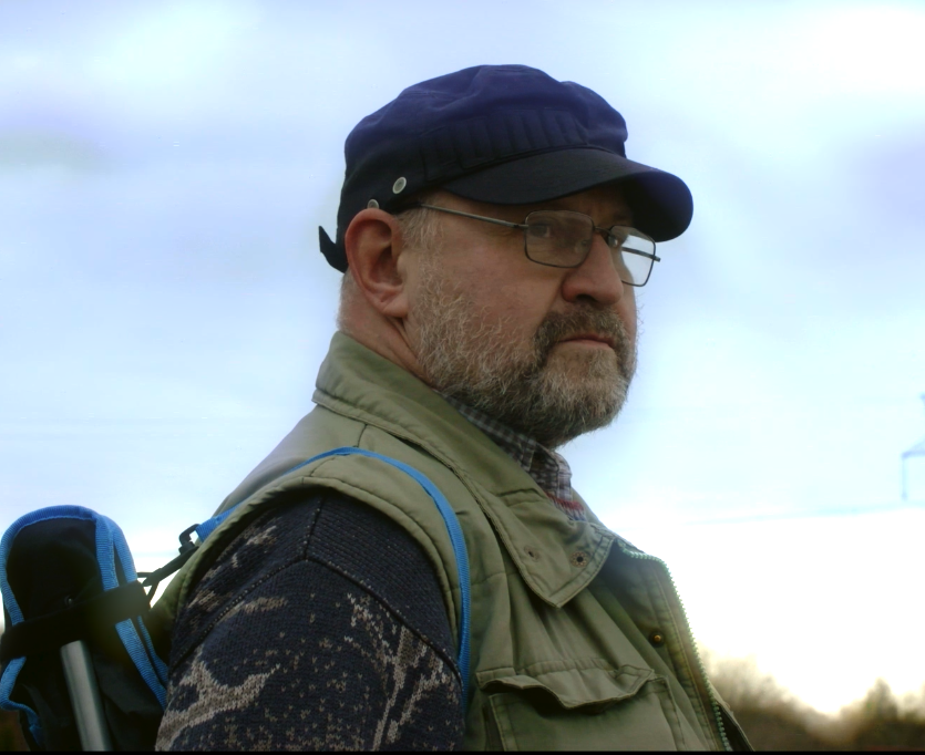 A man with glasses, a beard, wearing a navy cap and outdoor gear, standing outdoors with cloudy sky in the background.