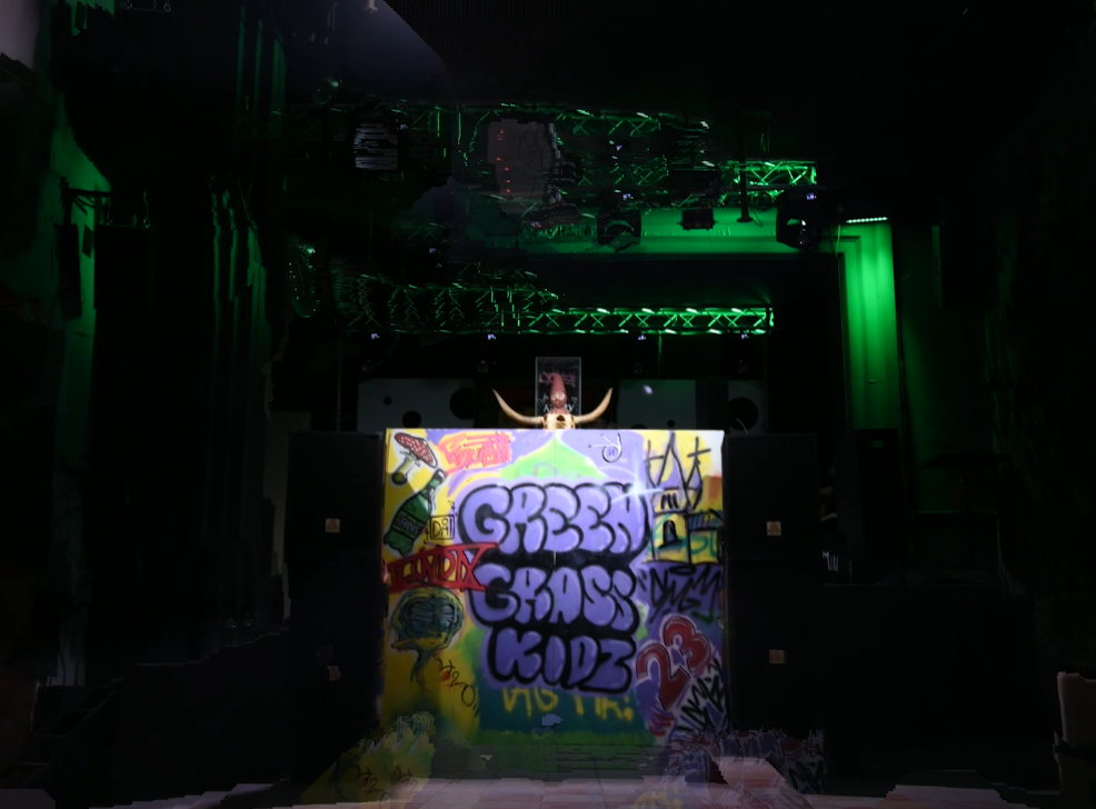 A DJ booth decorated with graffiti art, with a DJ wearing a hooded mask and horns, on a stage with green lighting and scaffolding overhead.