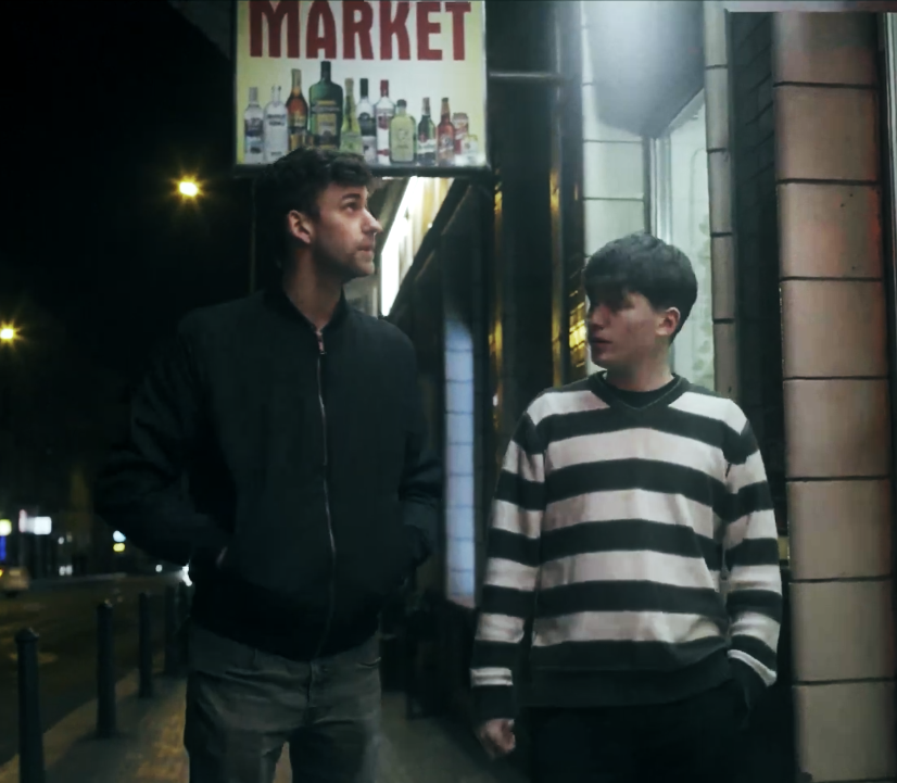 Two young men standing outside a store at night, with a sign above them that reads "Market" and shows images of alcohol bottles. One man is wearing a black jacket and the other a black and white striped sweatshirt.