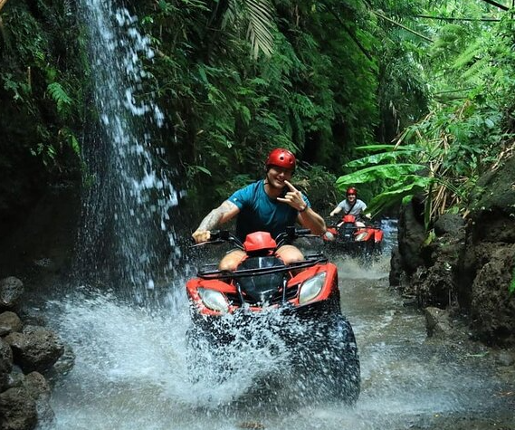 Two people riding all-terrain vehicles through a water stream in a lush jungle, both wearing helmets.