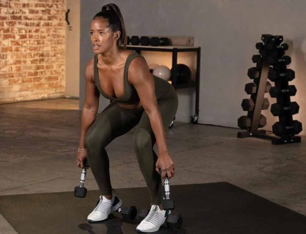 A woman in workout attire is performing a squat exercise with dumbbells in a gym.