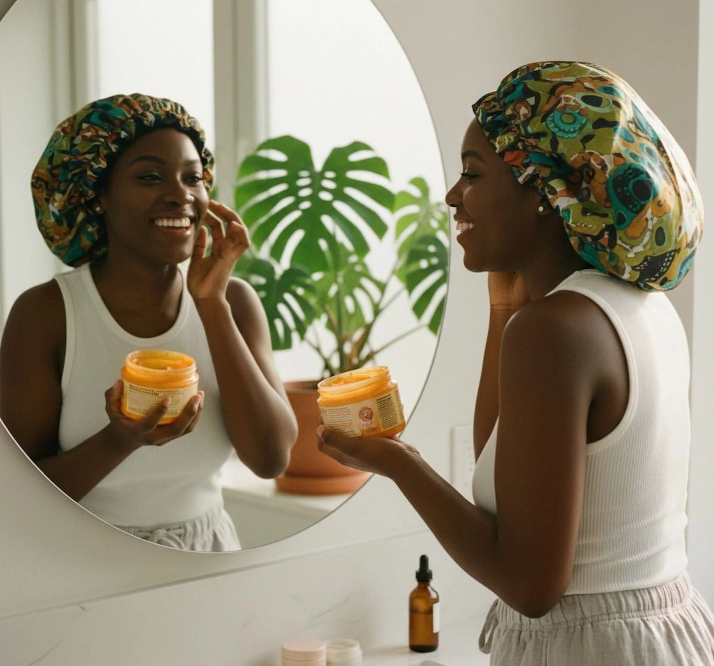 Woman wearing a colorful headscarf applying moisturizer from a jar while looking in a mirror, with skincare products on a bathroom counter and a plant in the background.