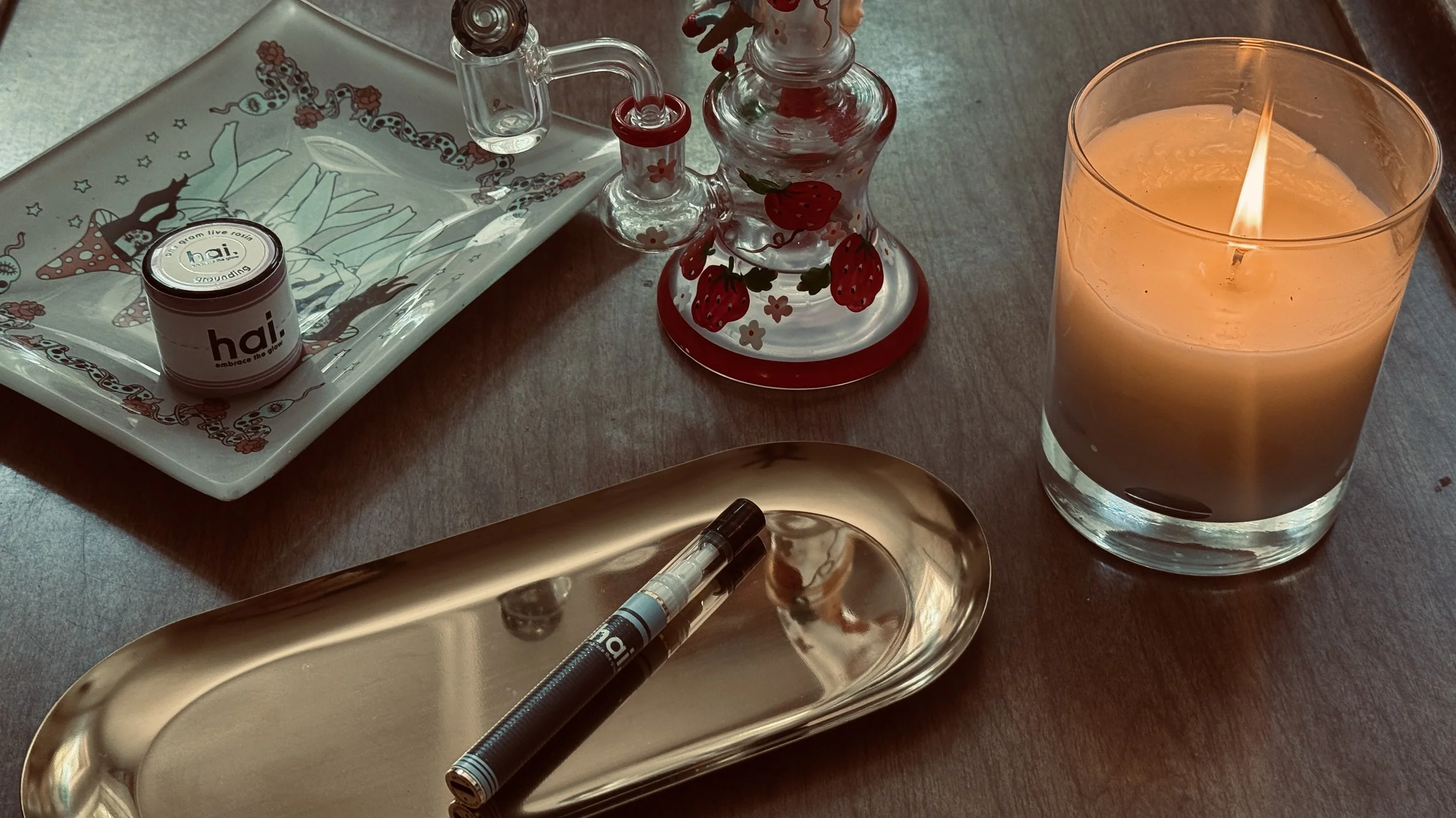hai. live rosin jar and vape pictured alongside a strawberry-adorned glass piece, illustrated tray, and lit candle on a warm wooden table, creating a cozy cannabis ritual setting.