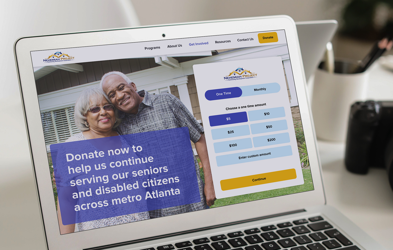 Laptop screen showing a donation webpage for Nehemiah Project, with an image of two smiling elderly people in front of a house, and options to donate one time or monthly, with amounts from five to two hundred dollars.