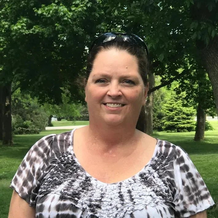 A smiling woman with sunglasses on her head standing outdoors in a park with green trees and grass