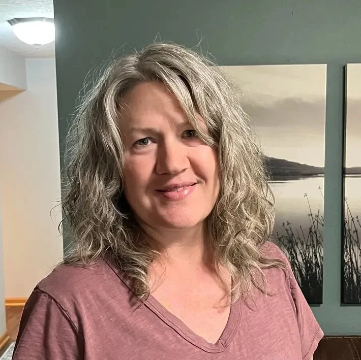 A woman with curly gray hair and a pinkish-purple shirt standing indoors in front of a green wall with landscape paintings.