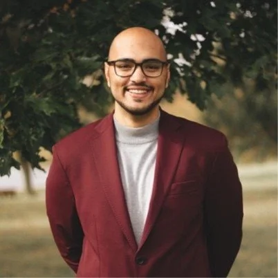 Smiling man with glasses, beard, and a bald head standing outdoors in front of a tree, wearing a maroon blazer and gray sweater