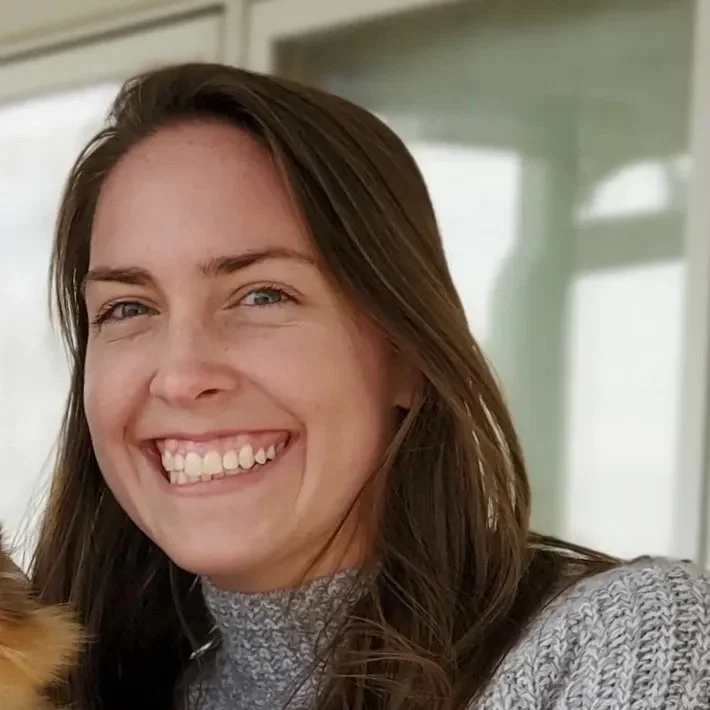 A woman with long brown hair, blue eyes, and a bright smile wearing a gray sweater, indoors with a window behind her.