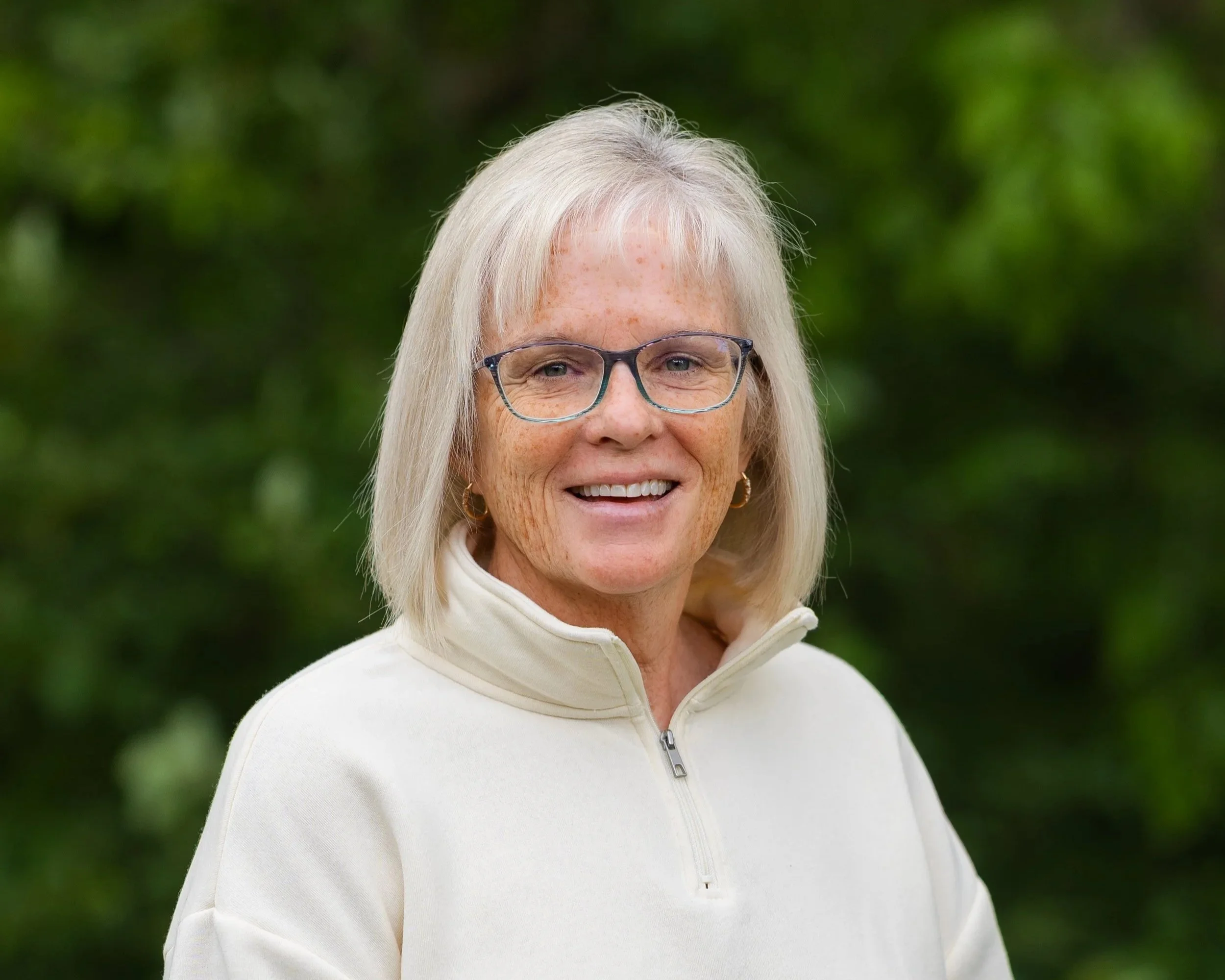 A smiling woman with blonde hair, glasses, and a red sports jacket, standing against a blue background.