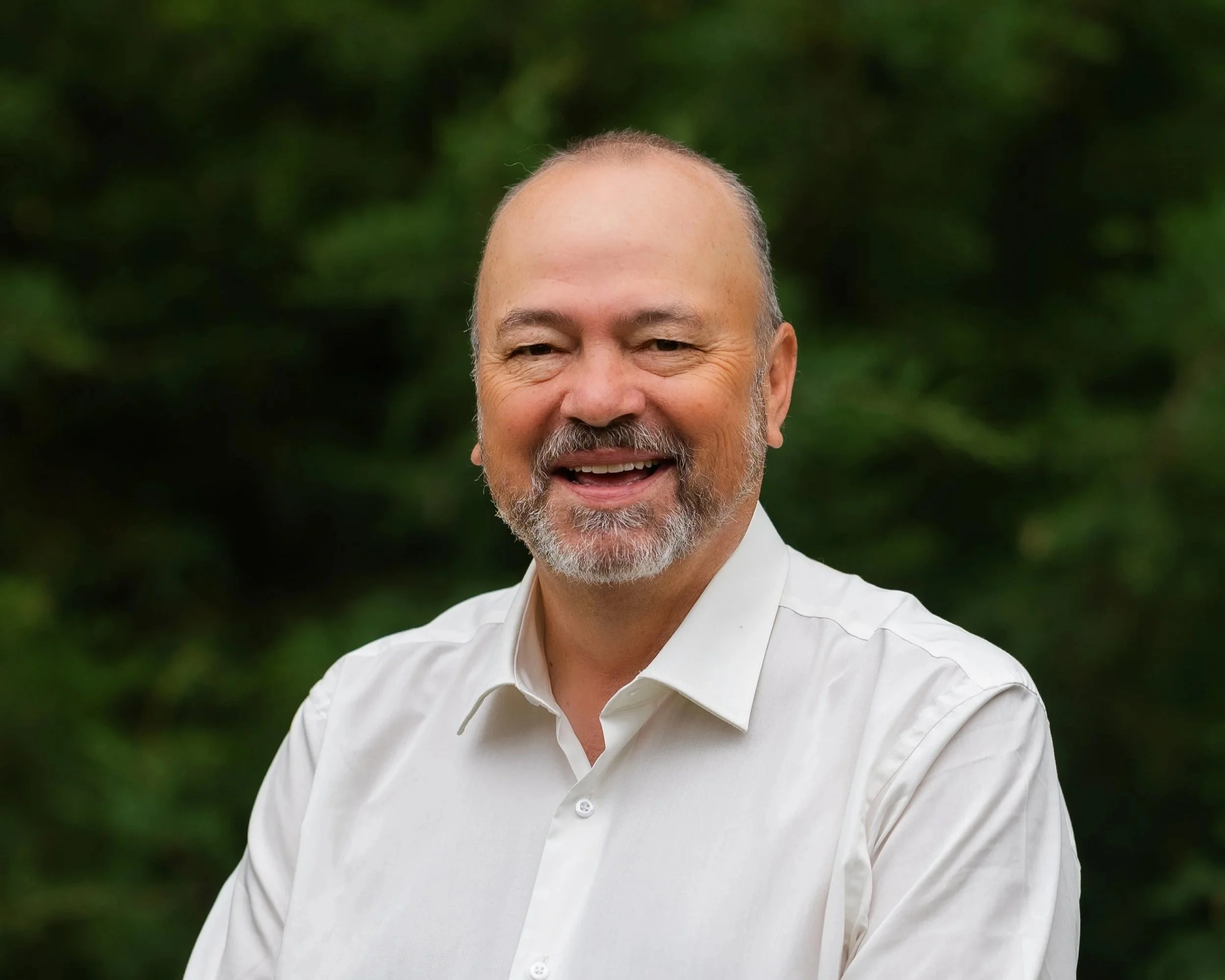 A smiling middle-aged man with a beard, wearing a navy suit, white shirt, and brown tie, with a boutonniere on his lapel, standing outdoors near a white column.