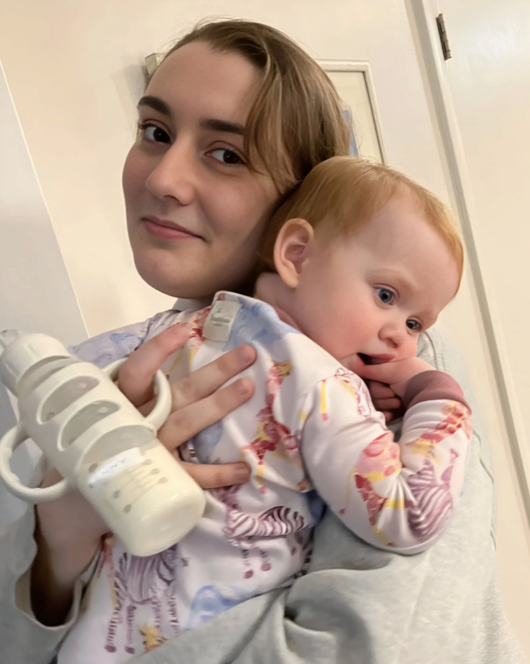 A young woman holding a red-haired toddler in her arms indoors. The toddler is resting with his head on her shoulder, holding a baby bottle, and appears to be sleepy or tired.