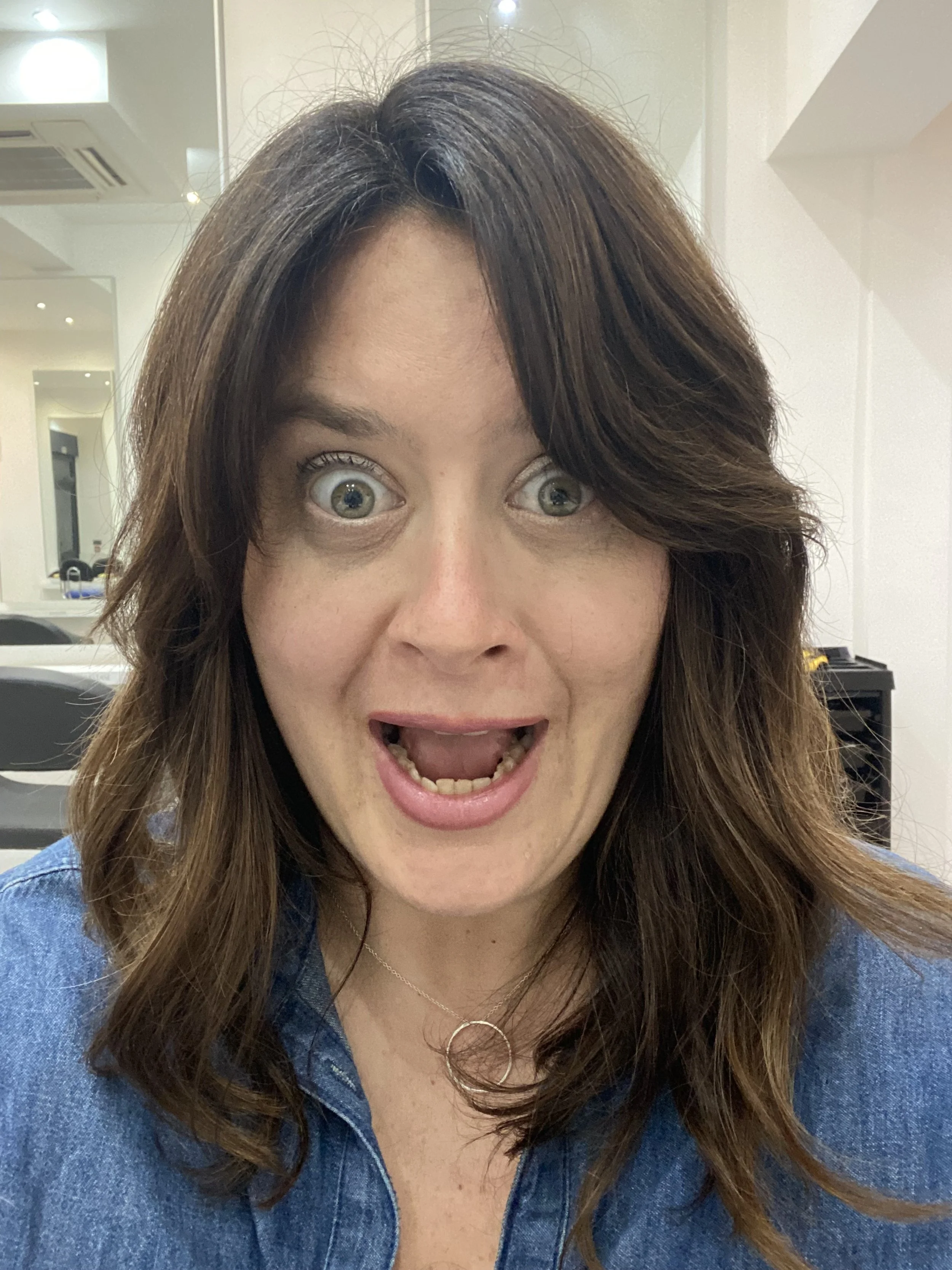 A woman with shoulder-length brown hair, blue eyes, and freckles, wearing a denim shirt and a necklace with two interlinked circles, making a surprised or excited facial expression.