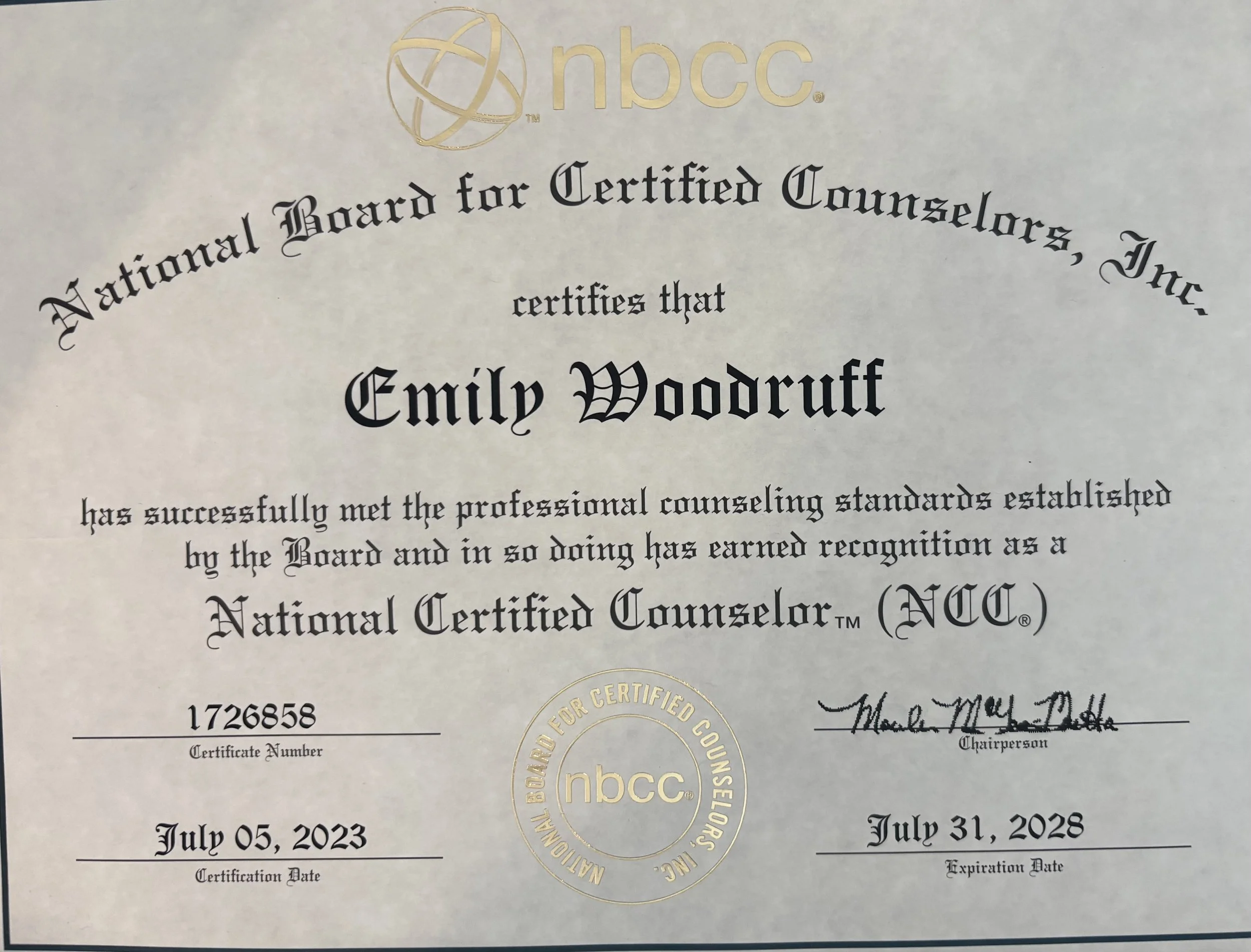 Close-up of a certificate from the National Board for Certified Counselors (NBCC), showing the recipient's name, Emily Woodruff, along with certification and expiration dates of July 05, 2023, and July 31, 2028 respectively, and signatures at the bottom.