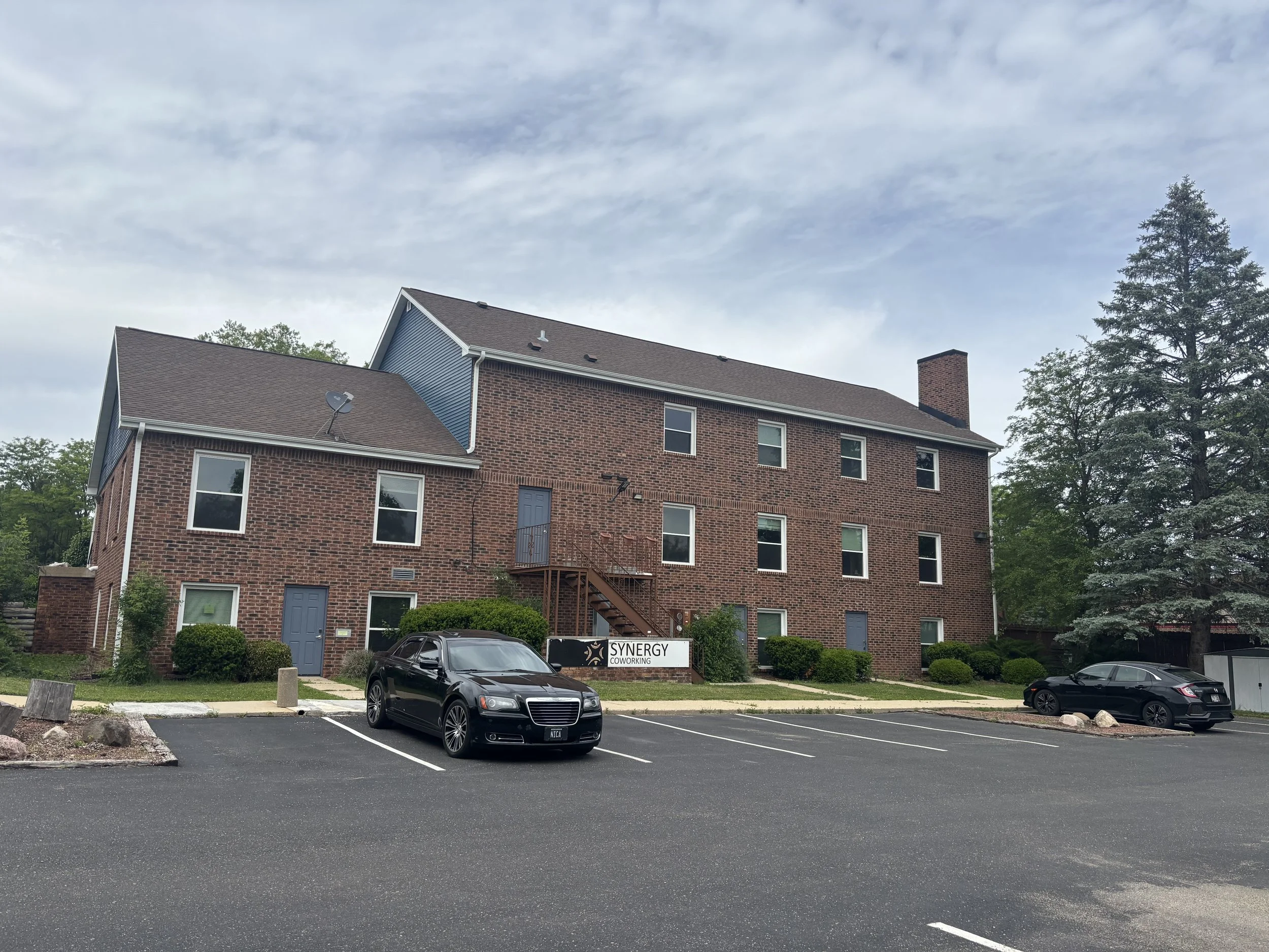 A brick office building with a parking lot in front and a sign that says 'Synergy Coworking'. There are two cars parked, a black one and a gray one. The sky is cloudy with some trees on the right side of the building.