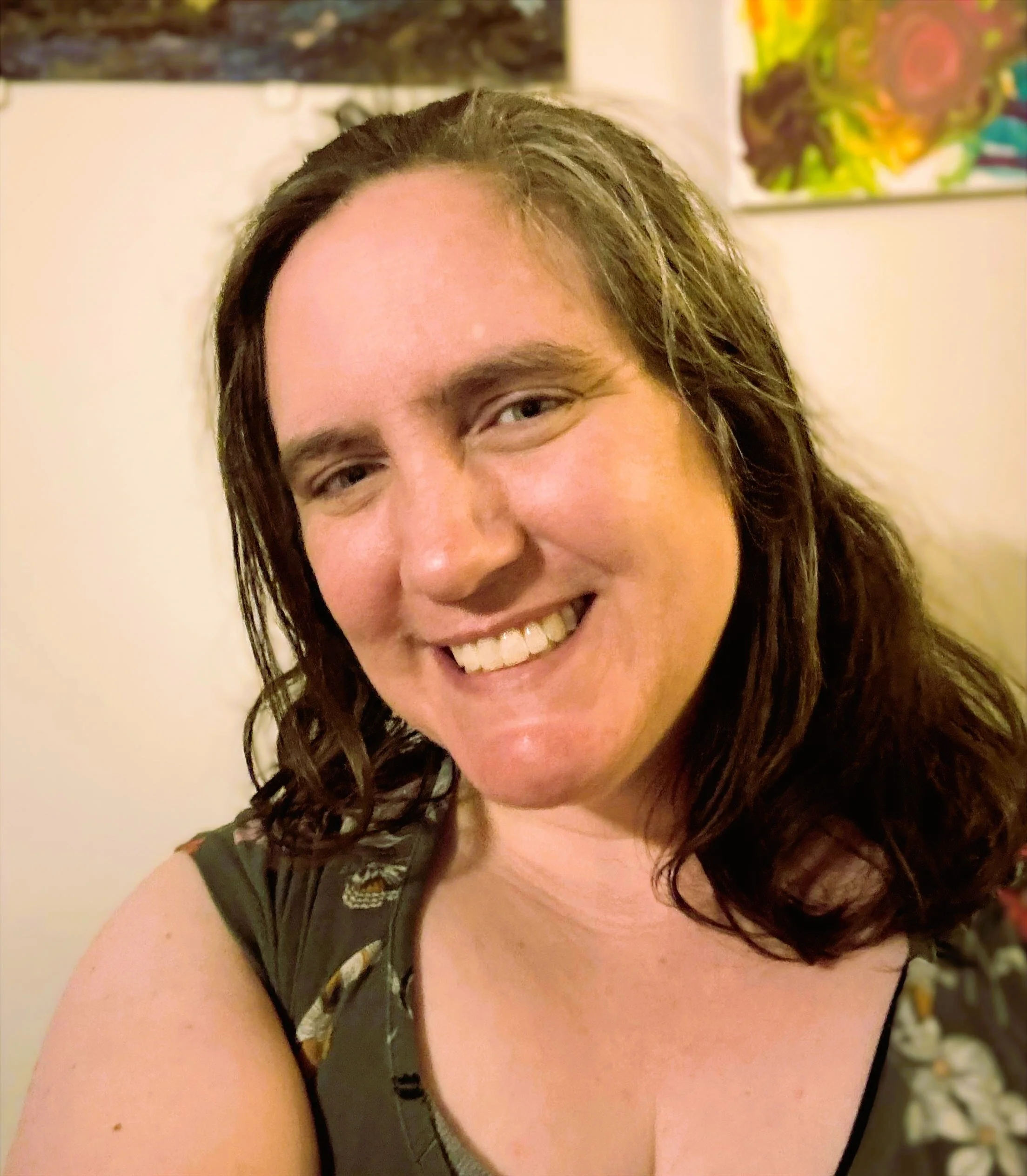 Close-up of a smiling woman with dark, wavy hair, wearing a black top, in an indoor setting with colorful artwork in the background.
