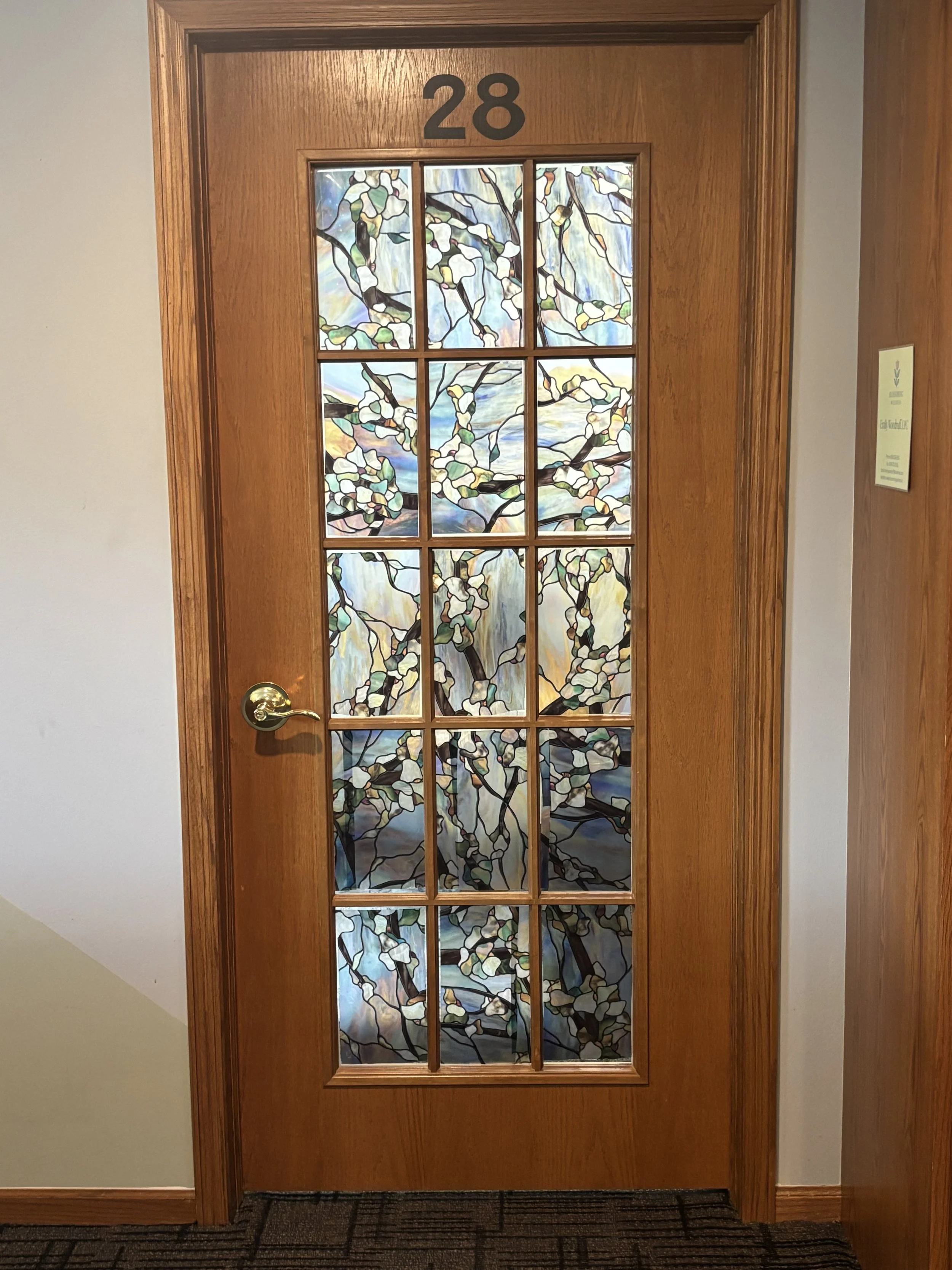 Wooden door to EMDR therapy office with glass panels featuring a stained glass design of tree branches and leaves, with a gold-colored door handle and the number 28 above the door.