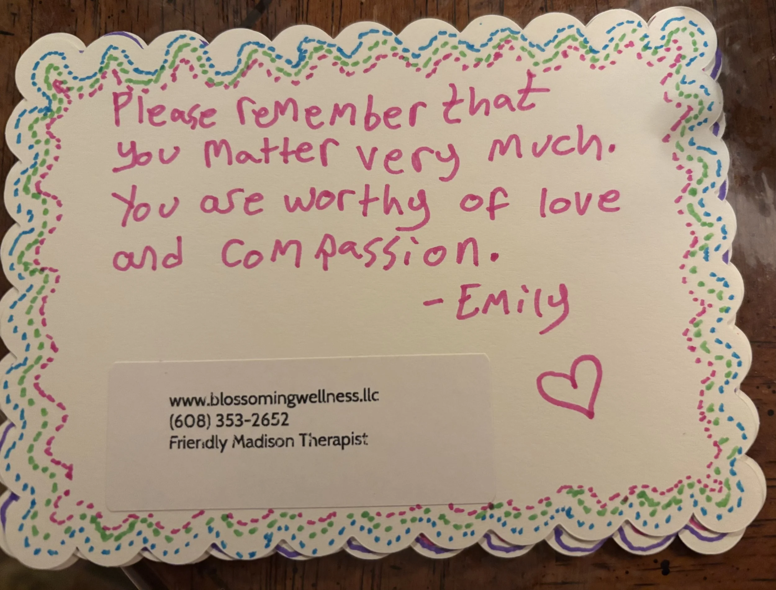 A decorative cut-out card with a colorful dotted border. It features a handwritten message in pink ink that says, 'Please remember that you matter very much. You are worthy of love and compassion.' The bottom of the card contains contact information for Blossom Wellness, including a website and phone number.