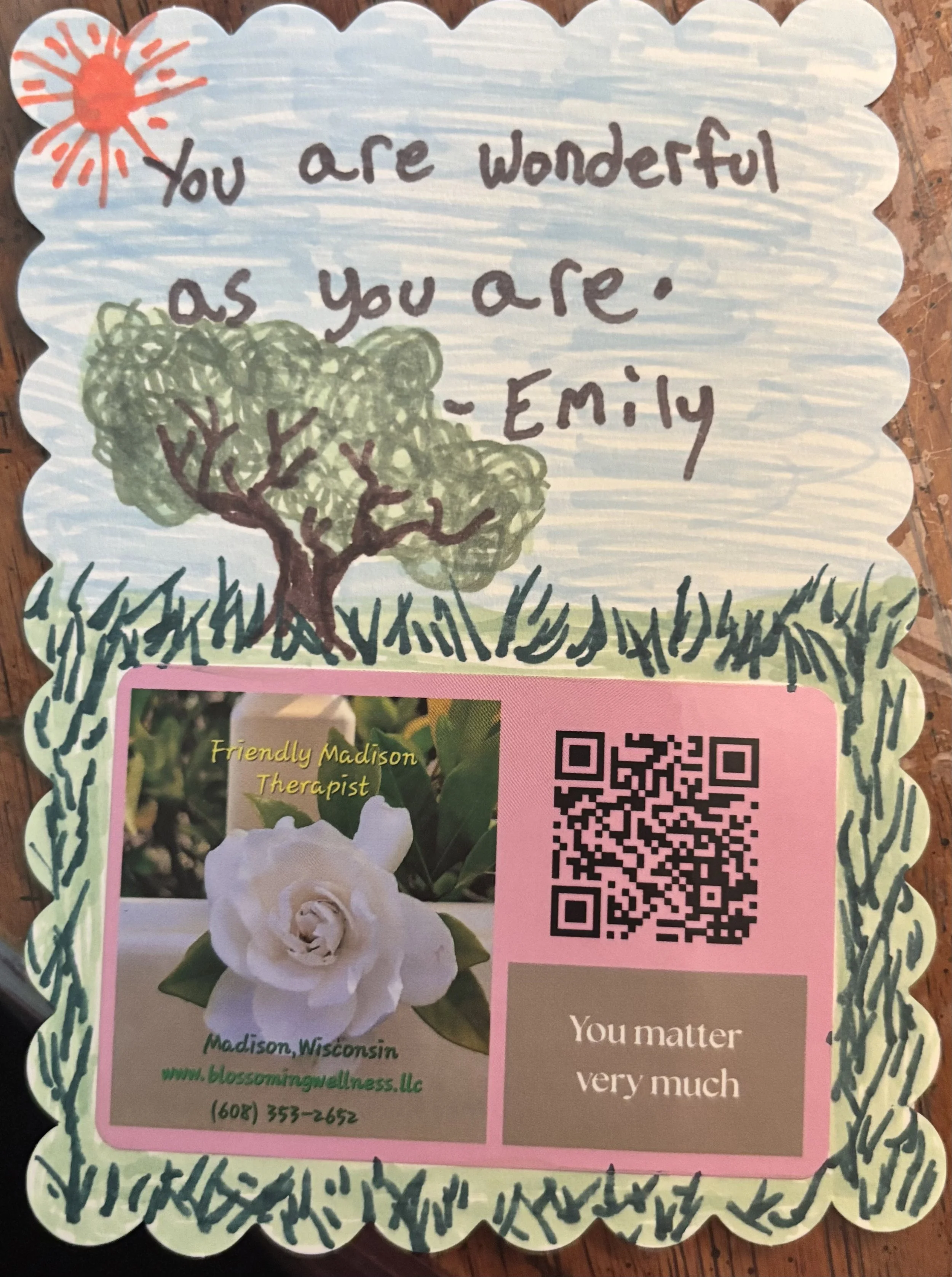 Hand-drawn illustration with a red sun, a green tree, and grass surrounding a white flower. Text above reads, 'You are wonderful as you are,' and additional text below appears on a business card that includes a photo of a white flower, contact information for Blossom in Wellness, LLC in Madison, Wisconsin, and a QR code.