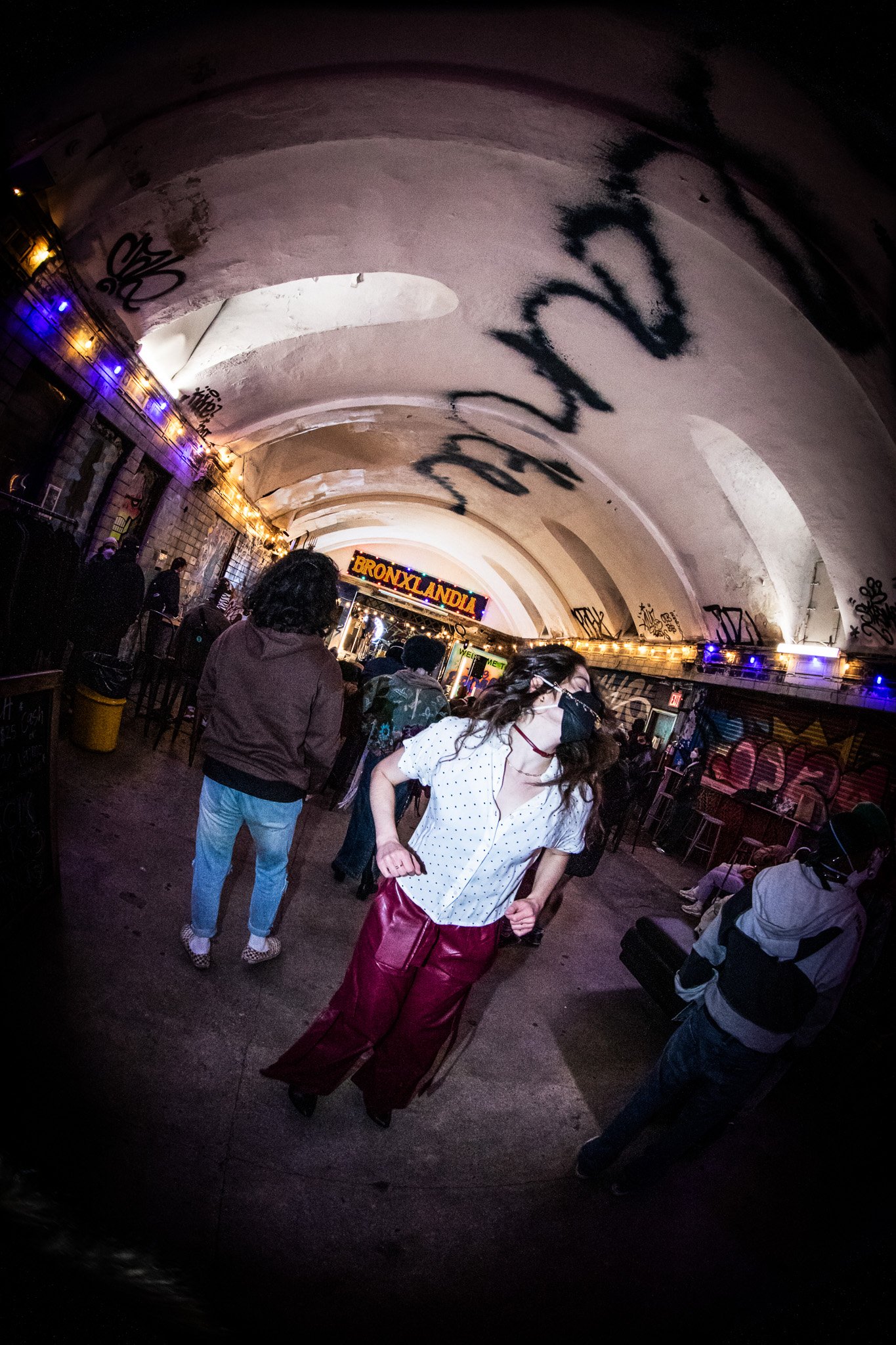 People dancing and talking at a bar with graffiti and colorful lights, with a sign reading 'BRONXLANDIA' in the background.