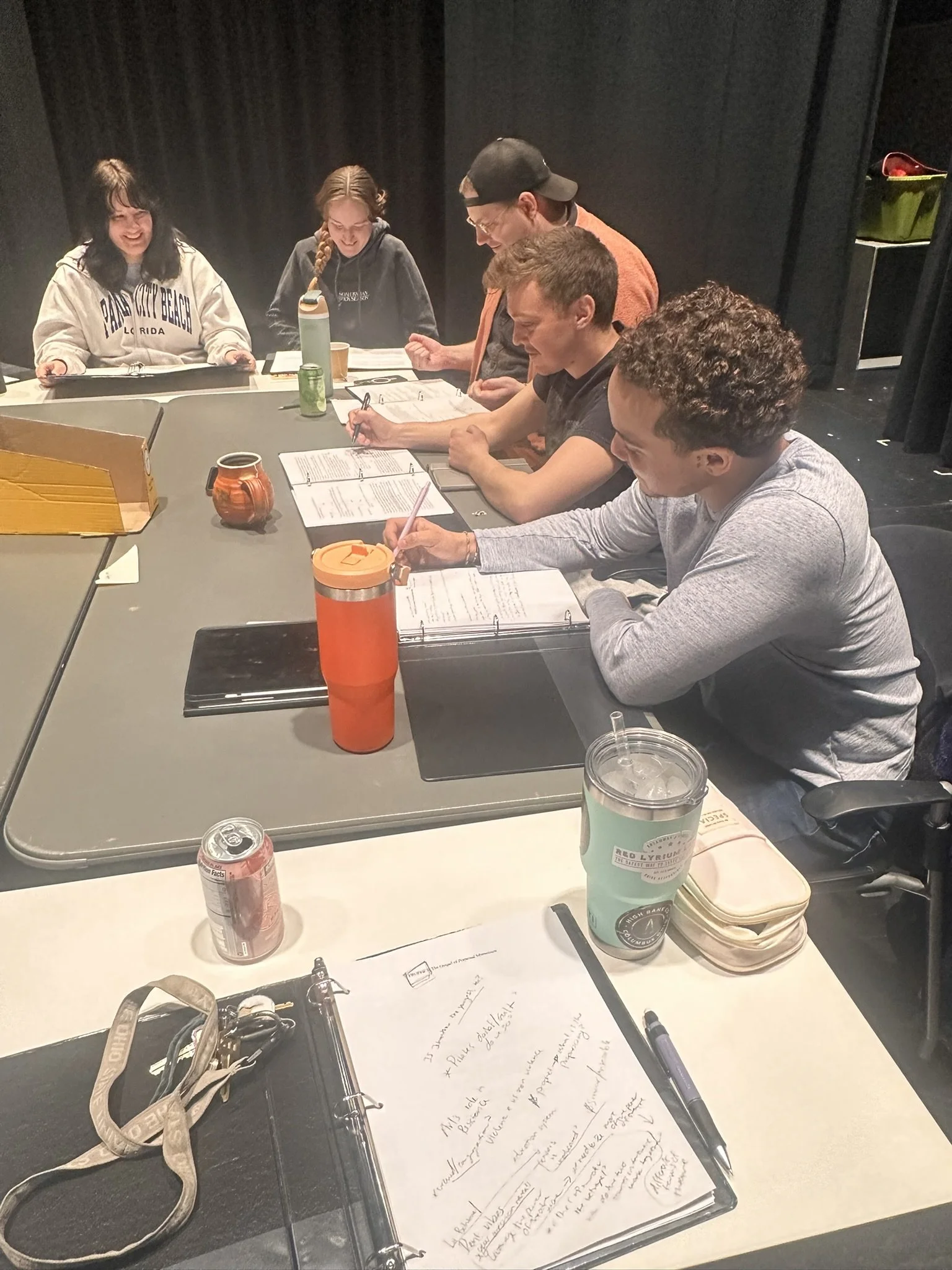 Group of six people sitting around a table, engaging in a meeting or discussion, with notebooks, drinks, and writing utensils on the table.