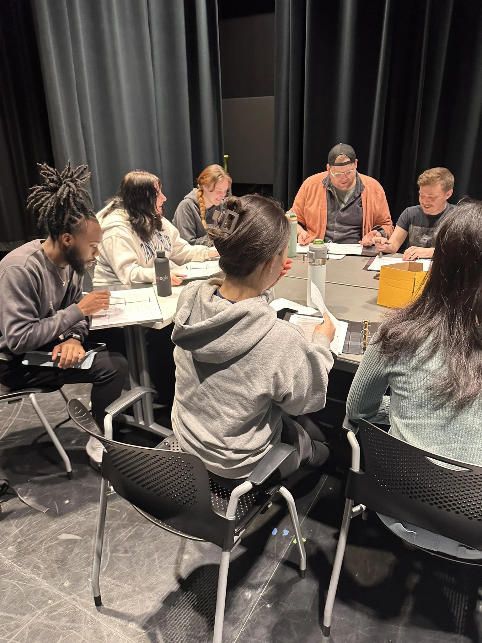 Group of diverse young people sitting around a table in a theater or classroom, engaged in discussion or studying.