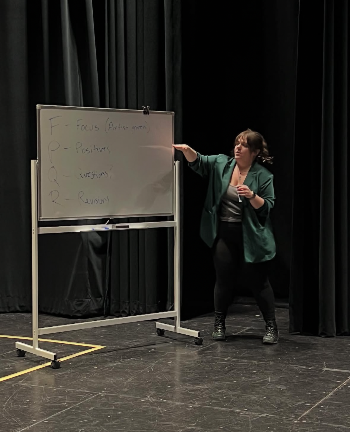 Megg stands on a stage next to a whiteboard with notes on it. She is wearing a green blazer and black pants, gesturing towards the whiteboard which has 'F-P-Q-R' written vertically with related terms.