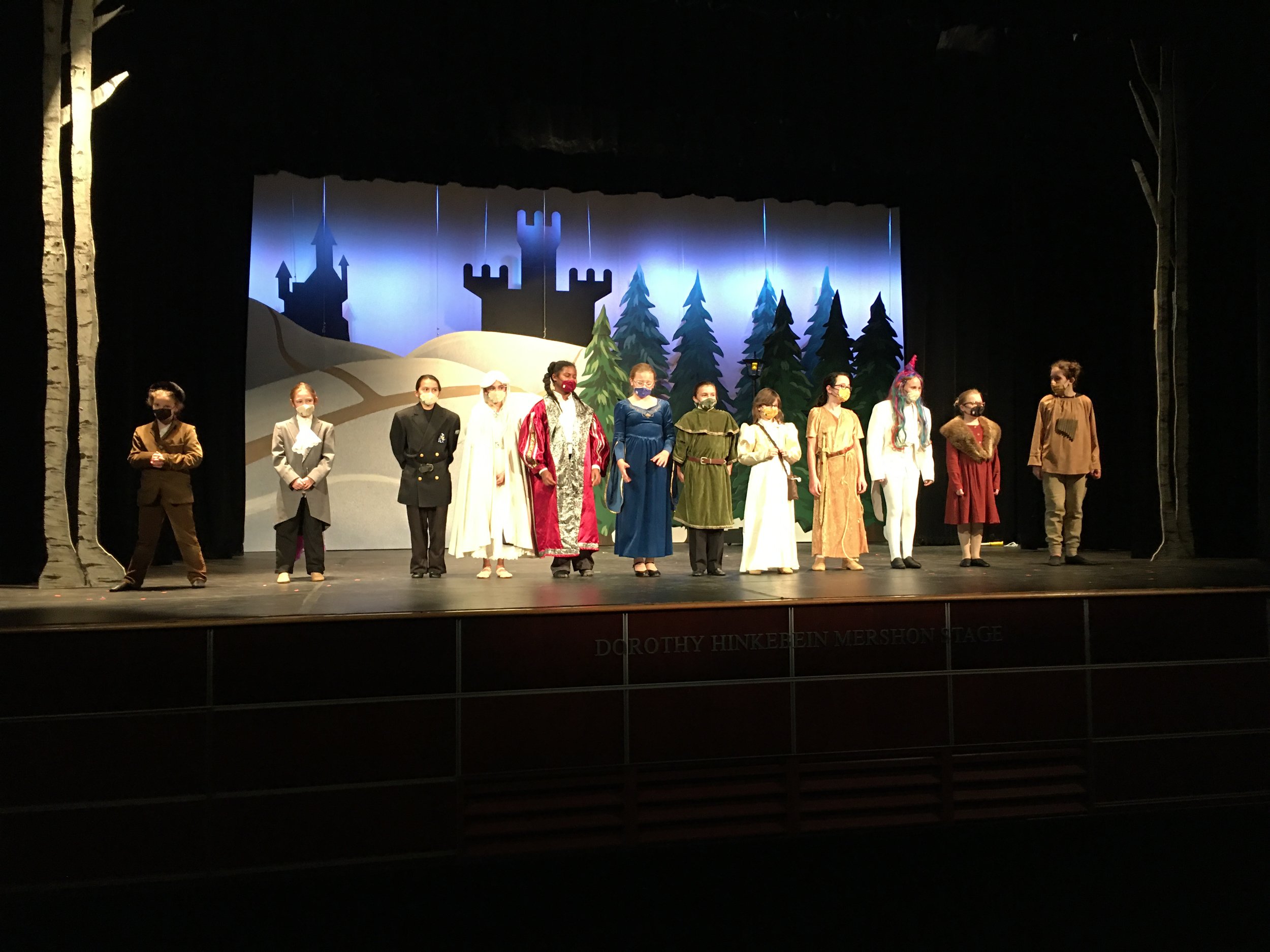 Children on stage in costumes for a play, with a backdrop of a castle, trees, and a snow-covered landscape, surrounded by black curtains.