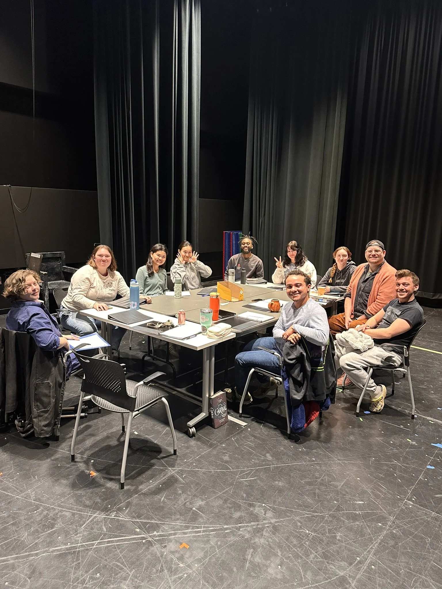 A group of eleven people sitting around a rectangular table on a stage, smiling at the camera.