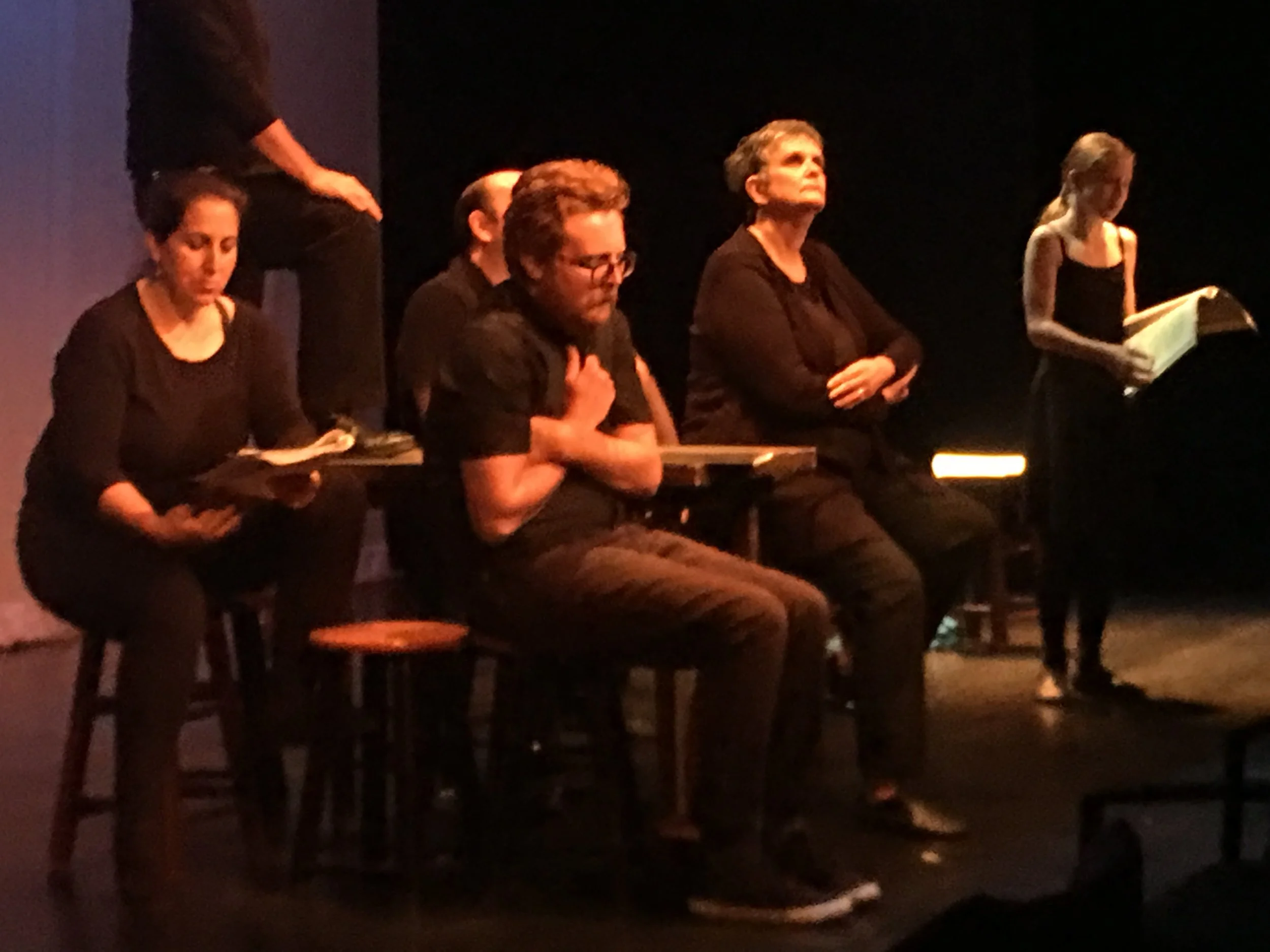 A group of people sitting on chairs on a stage, some reading or holding scripts, possibly rehearsing or performing a play.