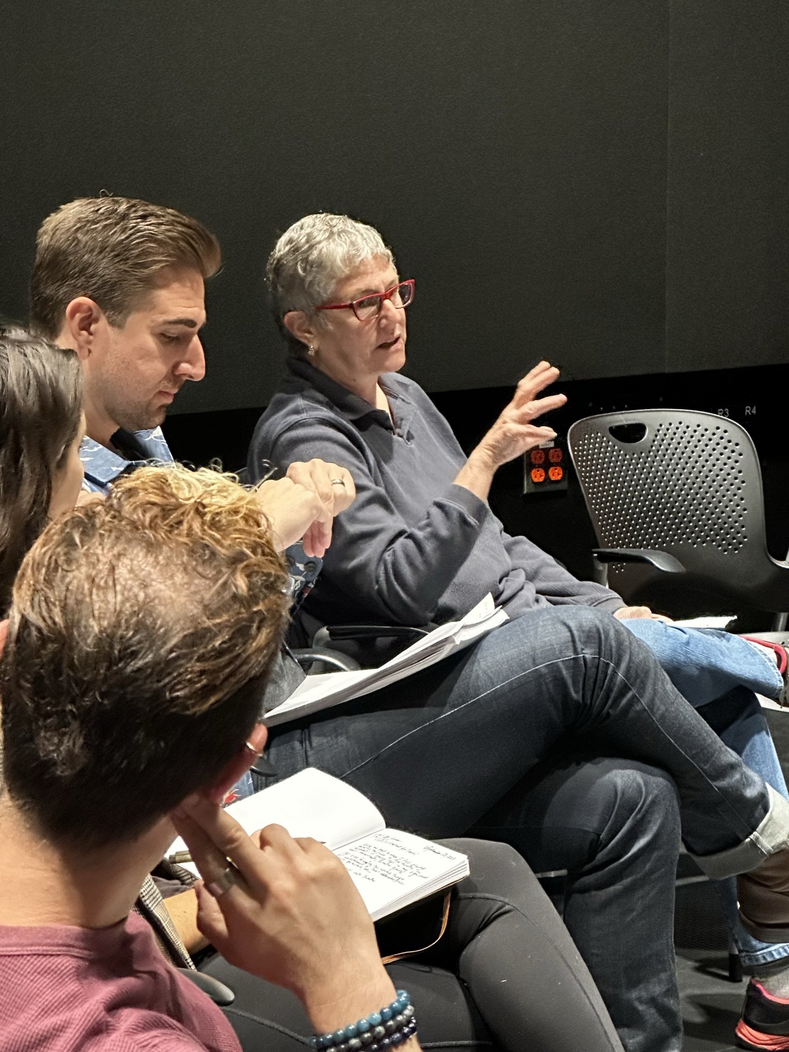 A group of people seated in a discussion or class, with a woman in glasses gesturing as she speaks.