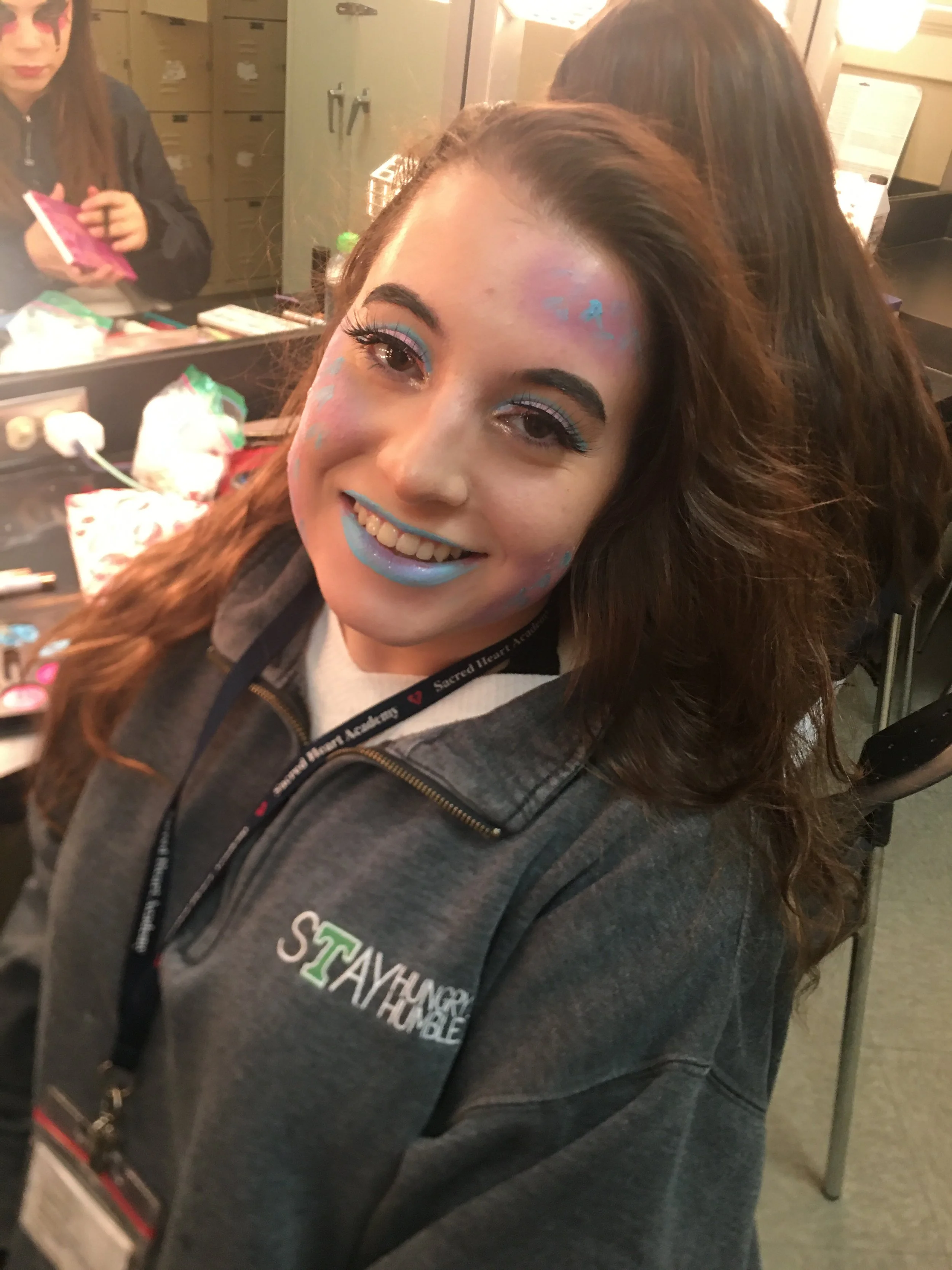 Young woman smiling with blue and pink face paint and makeup in an indoor setting, wearing a gray hoodie with 
