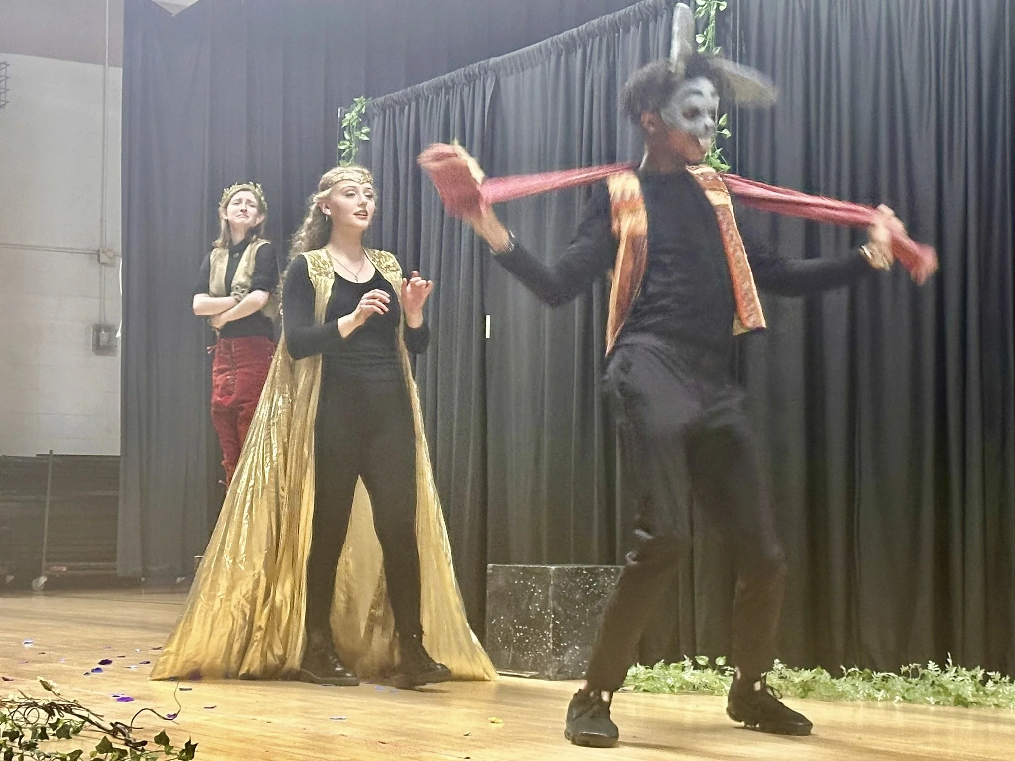 Three performers on stage, two women and one man, dressed in costumes for a theatrical performance with a black curtain backdrop. The man is wearing a mask and a costume with a mask, and is in a dynamic pose with a prop. The women are standing with o
