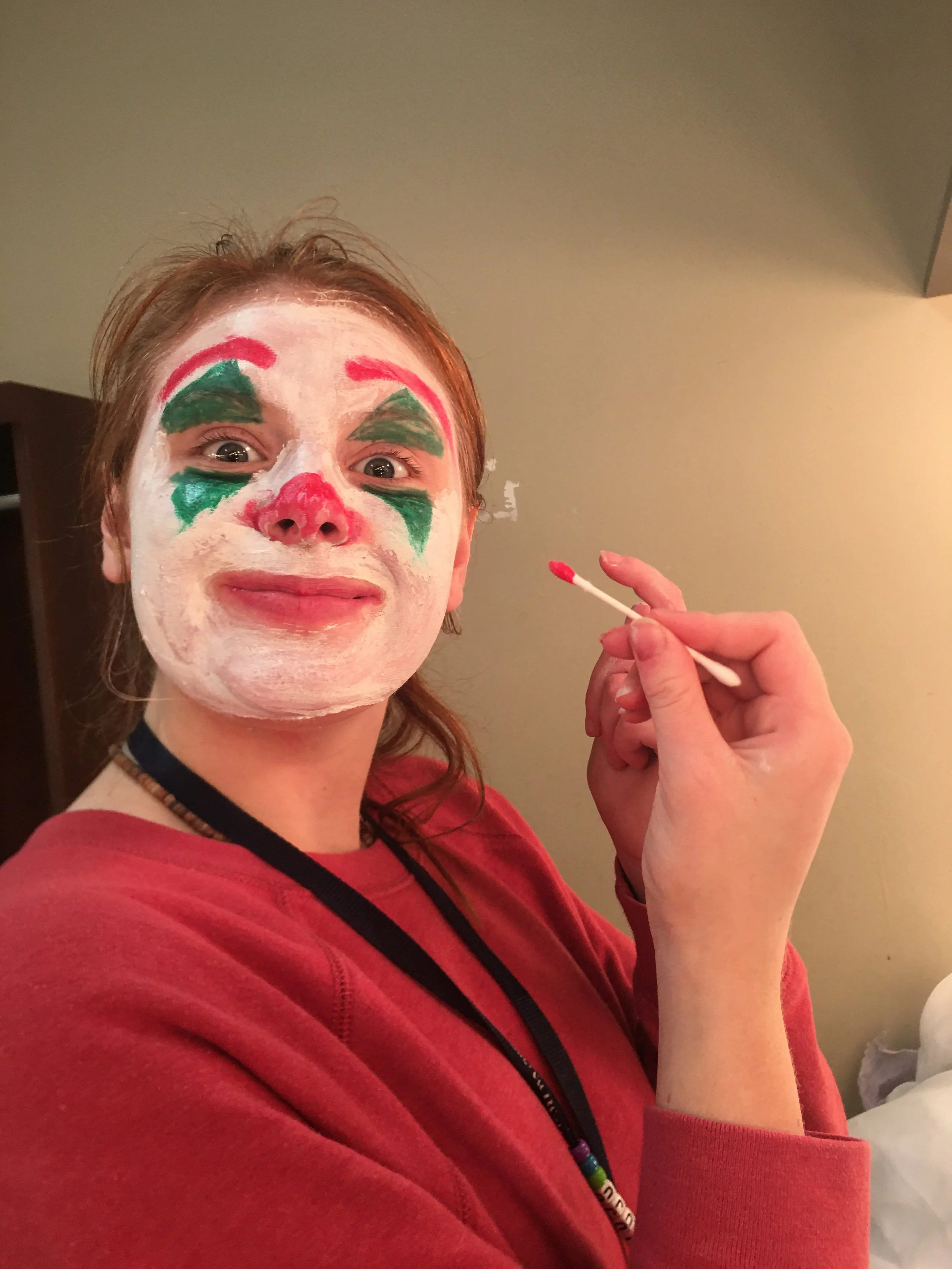 Person with face painted as a clown, white base, green eyebrows, red nose, and red lips, holding a cotton swab with pink paint.