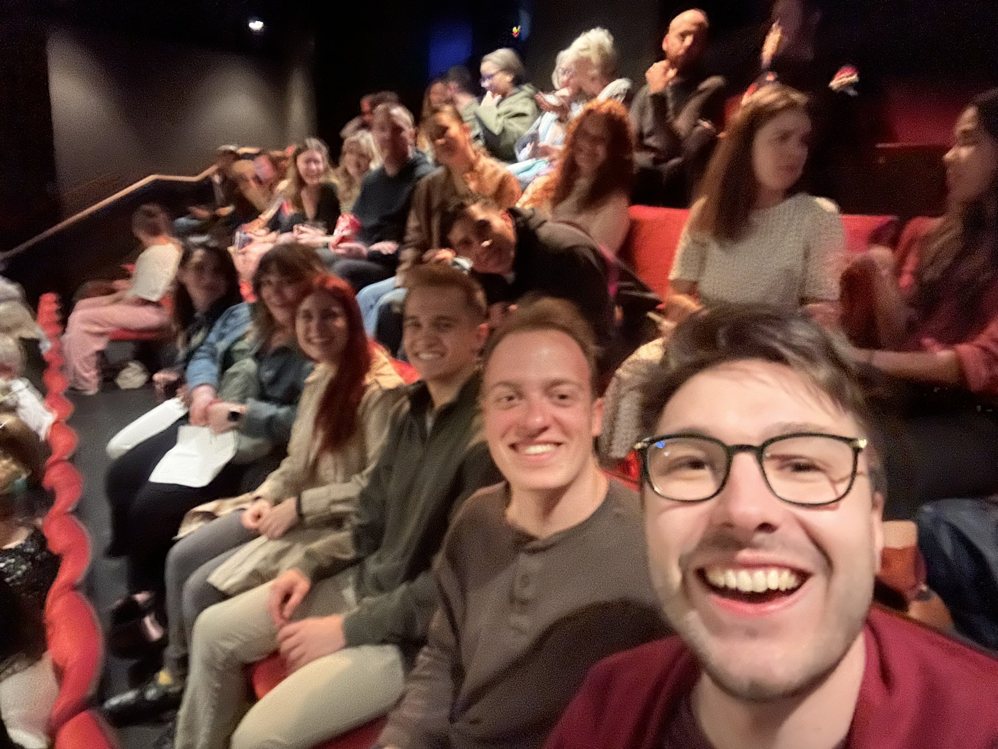 Group of people seated in a theater or cinema, taking a selfie. Many are smiling and looking at the camera, with some holding papers or programs.