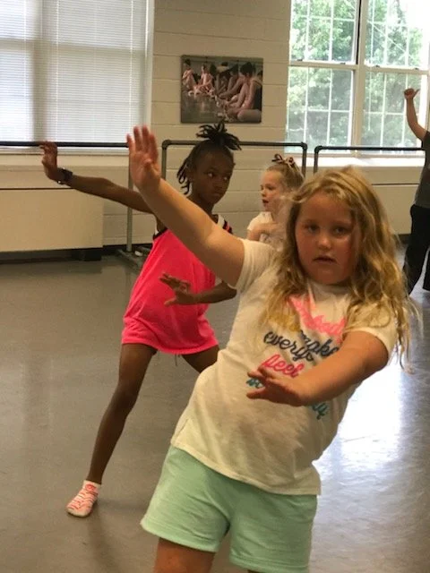 Young girls participating in a dance or movement class in a studio with large windows and ballet barre, with one girl in front extending her arms forward.
