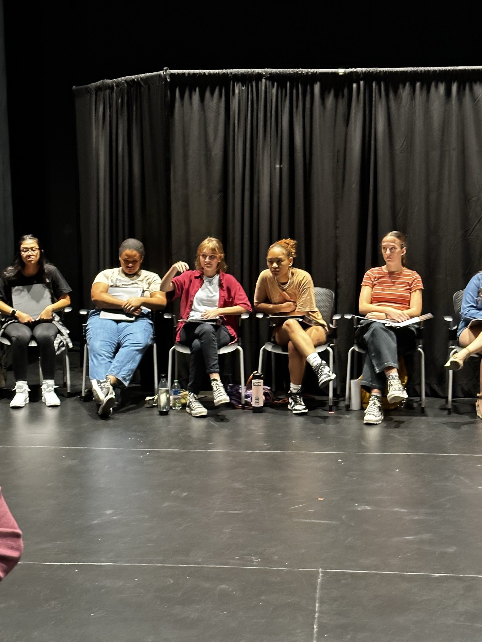 Six young women sitting in a row on stage chairs with a black curtain backdrop, appearing to participate in a panel or discussion.